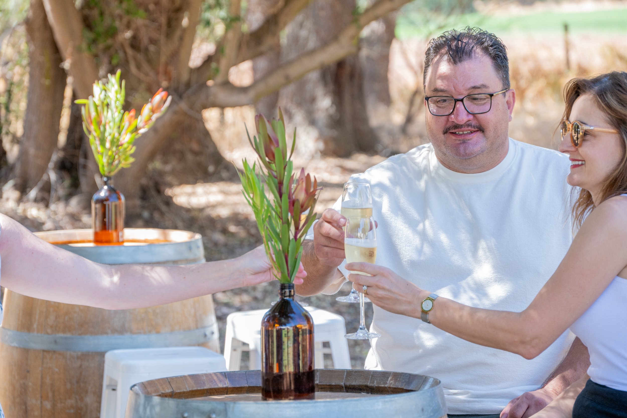 People outdoors at a gathering, one man wears glasses holding a glass of white wine, woman in sunglasses smiling, decorative bottles with flowers on barrels