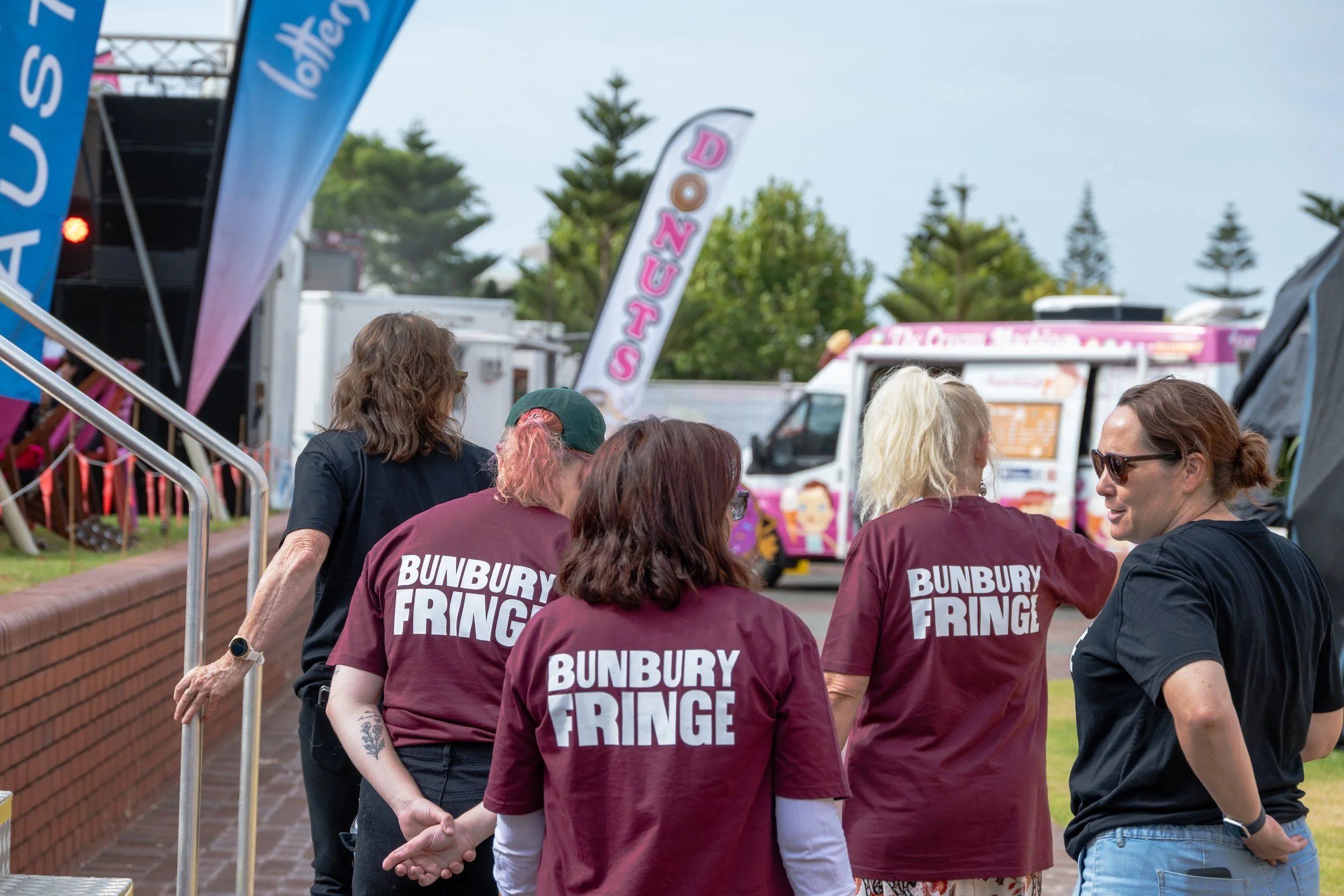 Group of five women standing outdoors near food vans and banners at an event, some wearing maroon shirts with 'BUNBURY FRINGE' printed on the back.