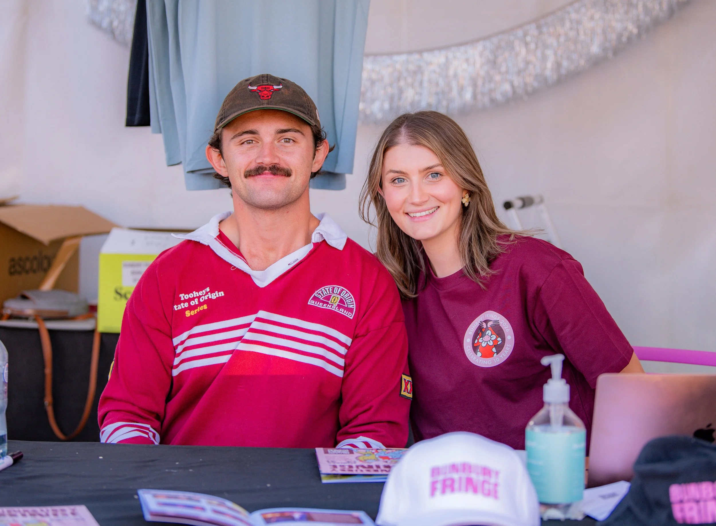 Two people sitting at a table, smiling at the camera, with various items including shirts, hats, and a bottle of hand sanitizer in front of them, inside a tent or booth.