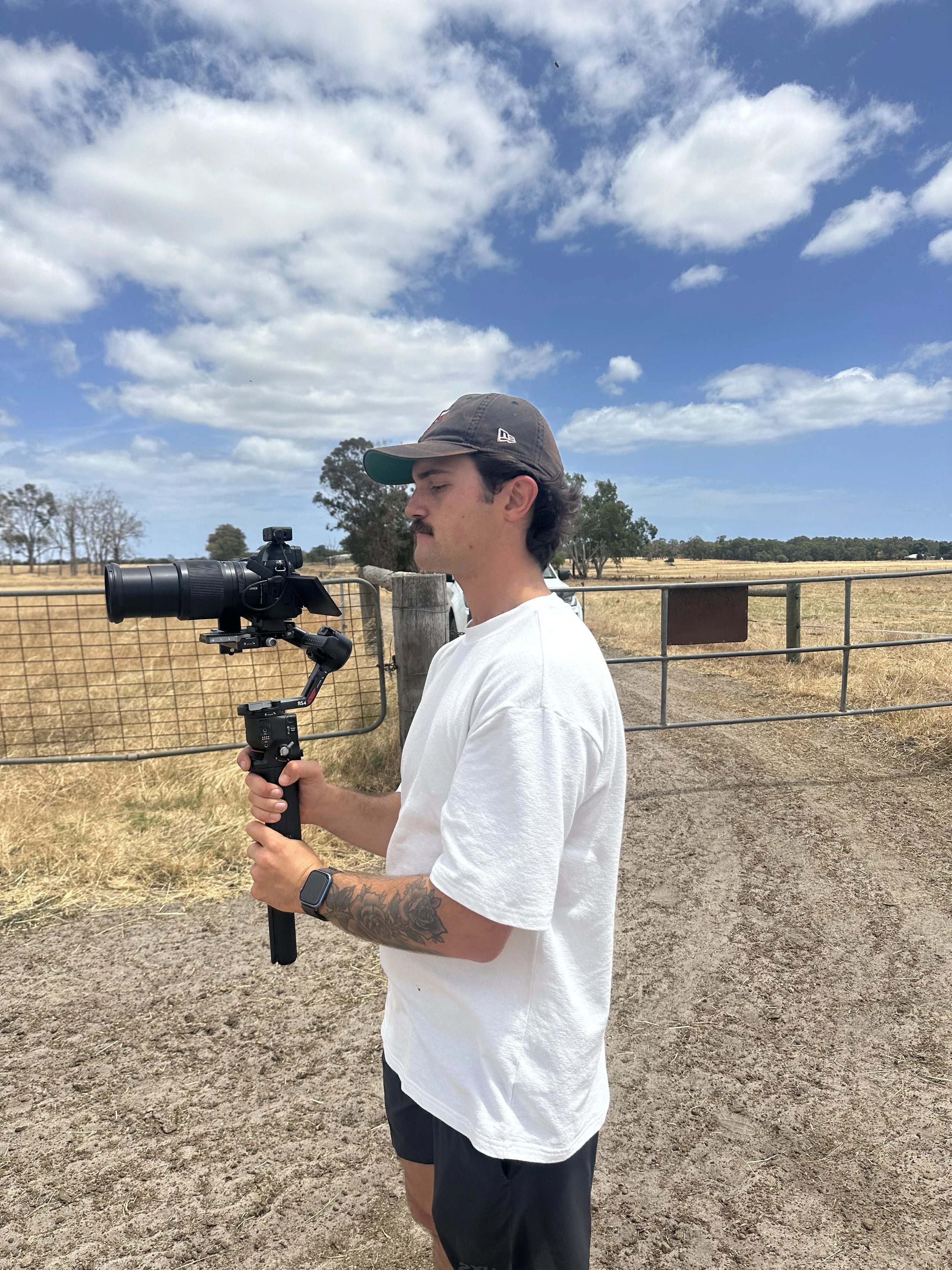 A young man in a white t-shirt, black shorts, and a baseball cap is holding a camera with a stabilizer on a dirt path near a farm gate in a rural area with trees and open fields under a blue sky with scattered clouds.