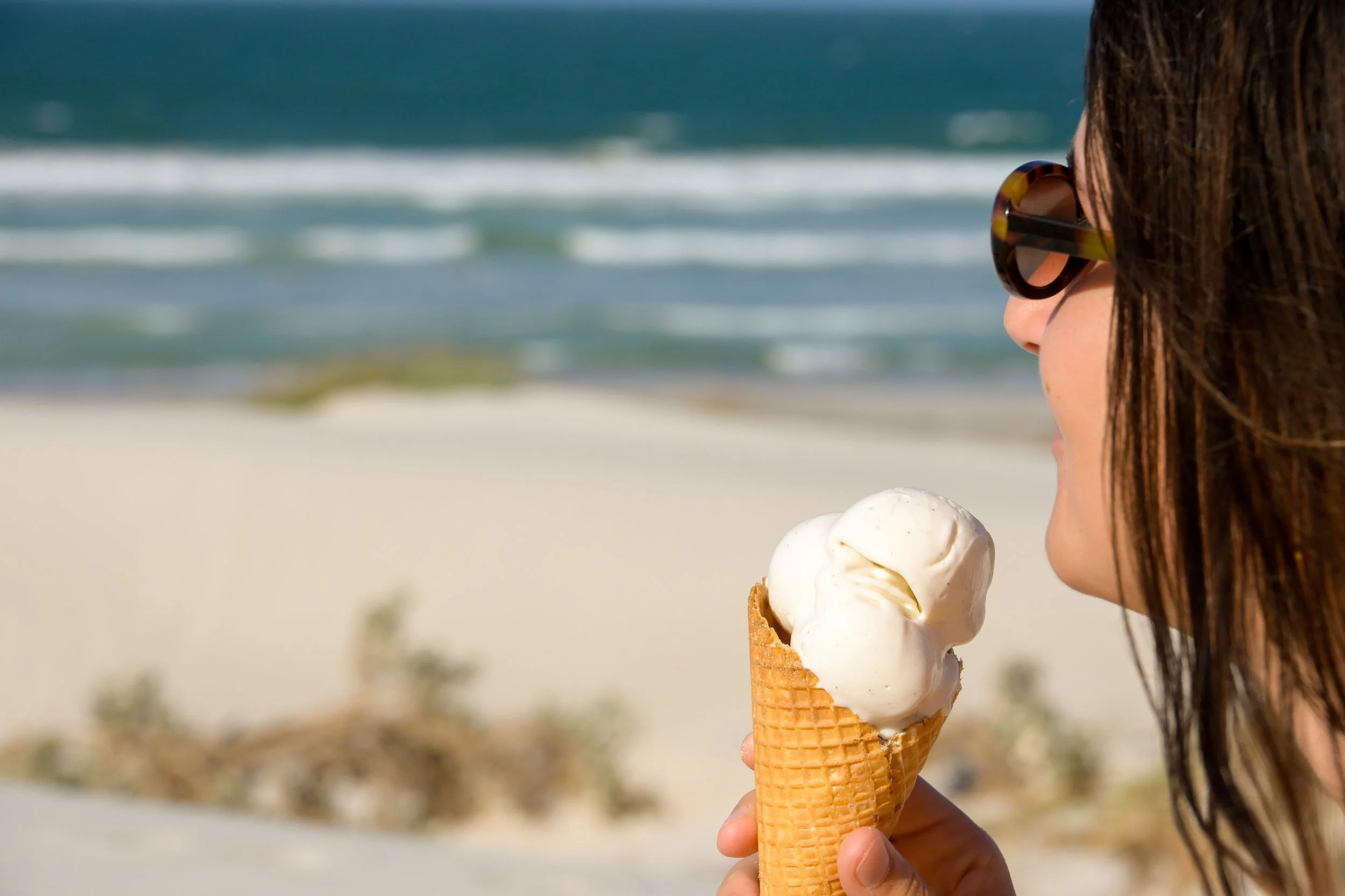 A woman with brown hair, wearing sunglasses, holding a vanilla ice cream cone on a beach with sand and the ocean in the background.