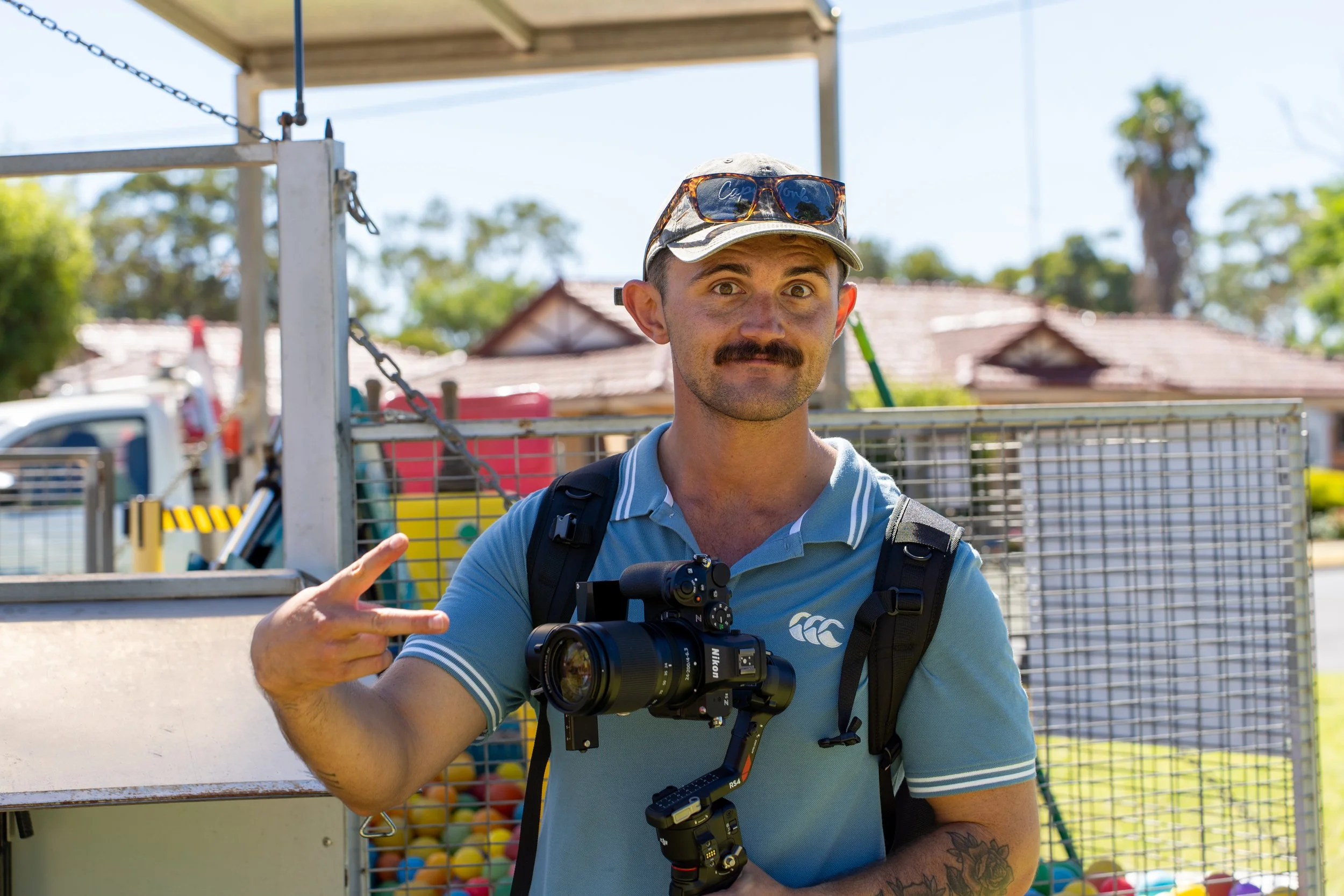 A man with a mustache wearing a light blue polo shirt, sunglasses, and a backpack holds a professional camera and makes a peace sign with his right hand.