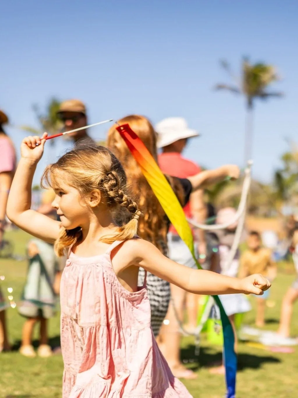 A young girl with braided hair wearing a pink dress is playing with a ribbon in an outdoor setting, surrounded by other children and adults, under a clear blue sky.
