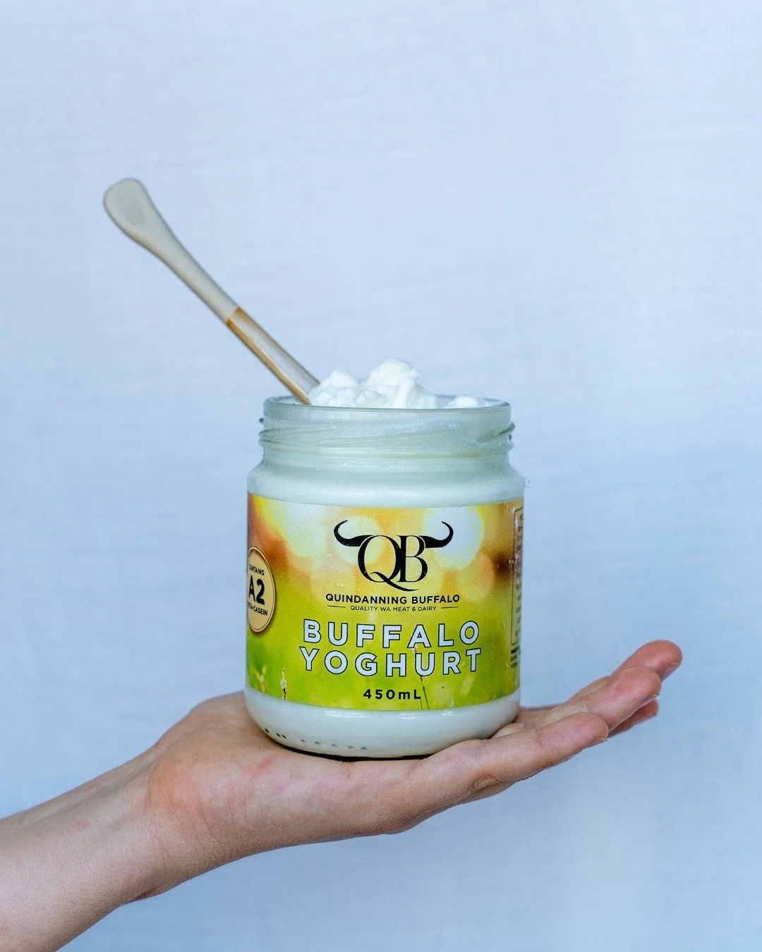 Hand holding a jar of Buffalo yoghurt with a wooden spoon inside, against a plain background.