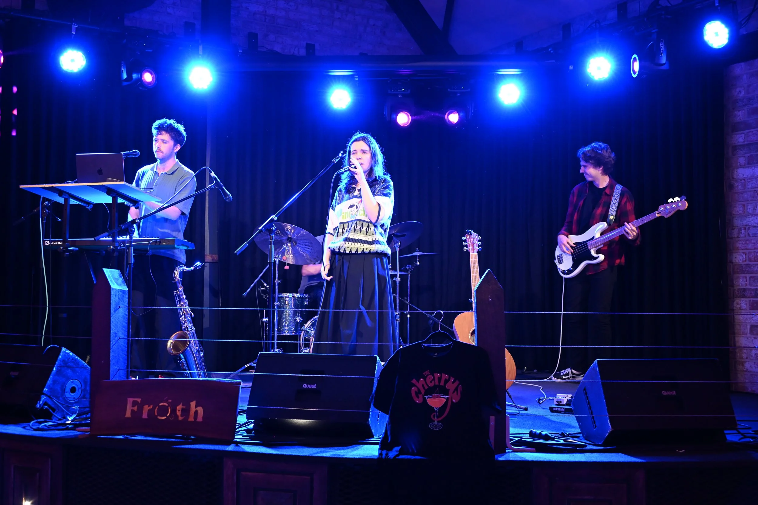A band performing live on stage with blue stage lights, including a keyboard player, a female vocalist, a drummer, and a bassist. The stage has a sign with 