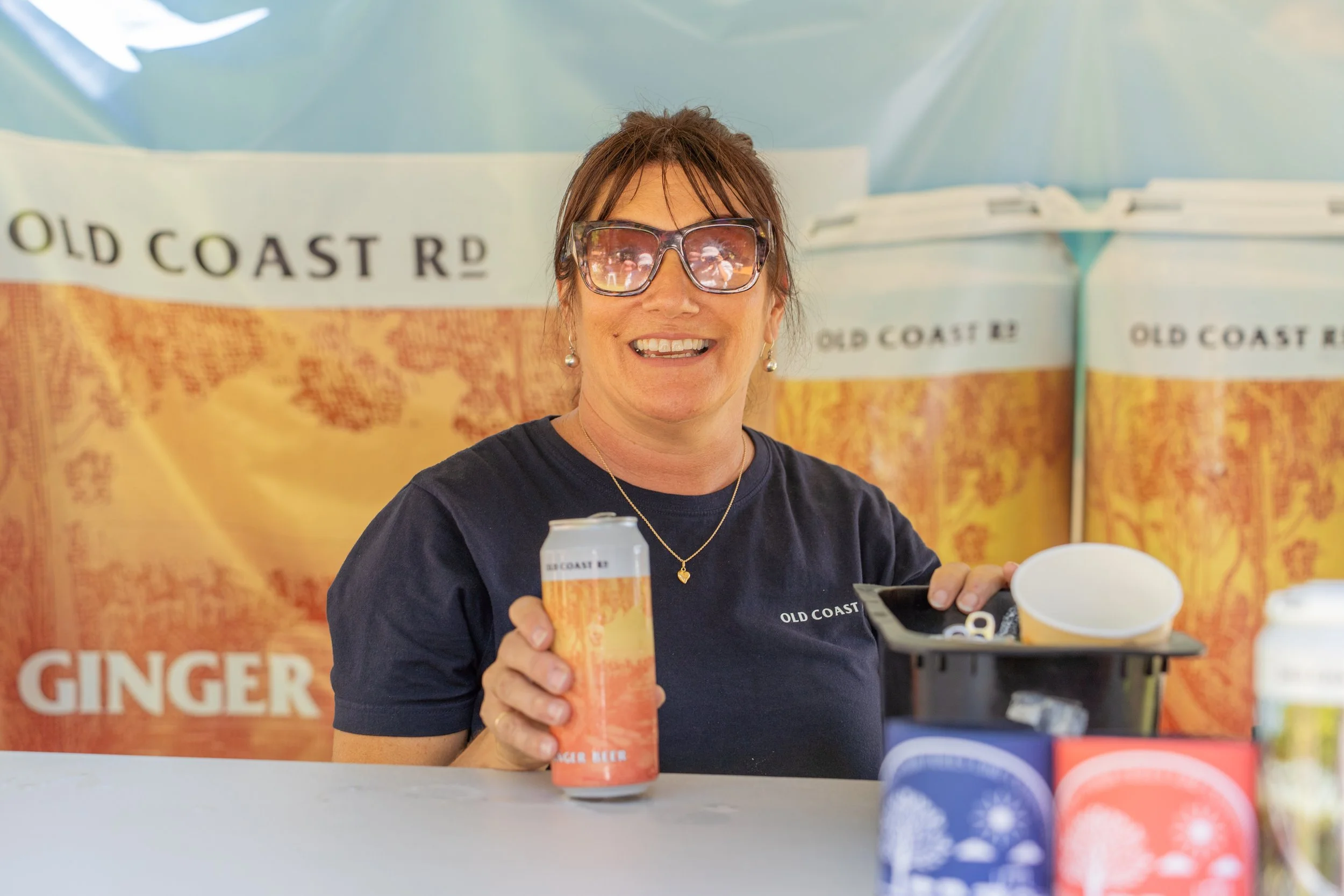 A woman with brown hair, wearing sunglasses and a black shirt, smiling and holding a drink can in front of a promotional background for Old Coast Ginger Beer.