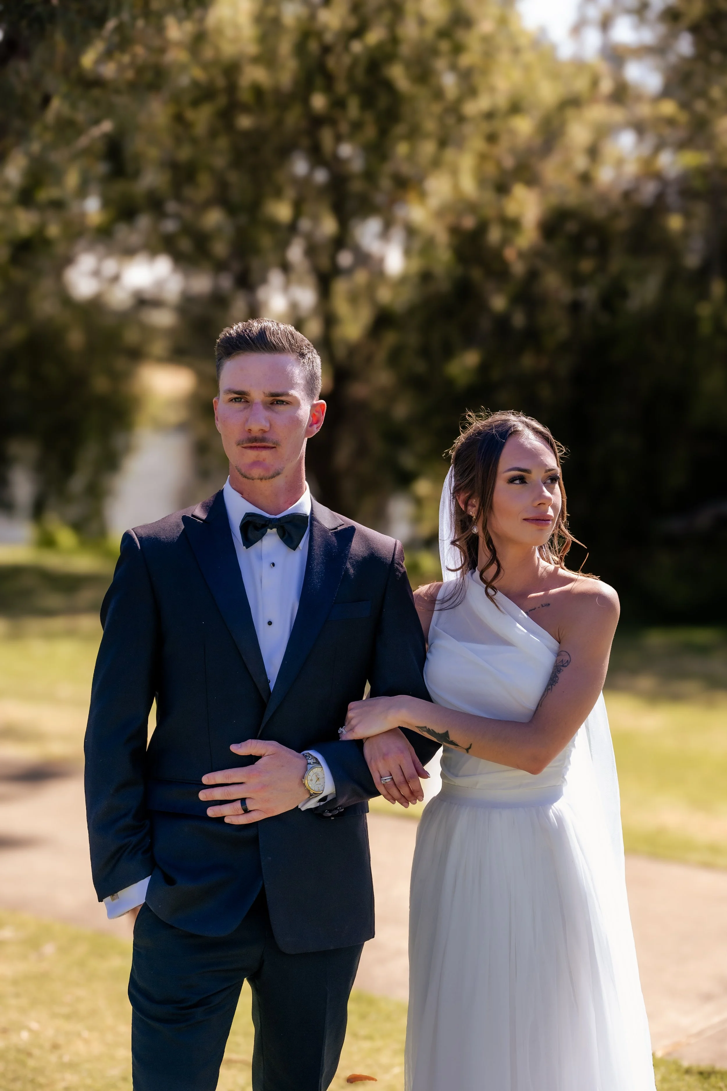 A bride and groom standing outdoors on their wedding day, with the groom in a black tuxedo and bow tie and the bride in a white gown with a veil, holding each other's arms.