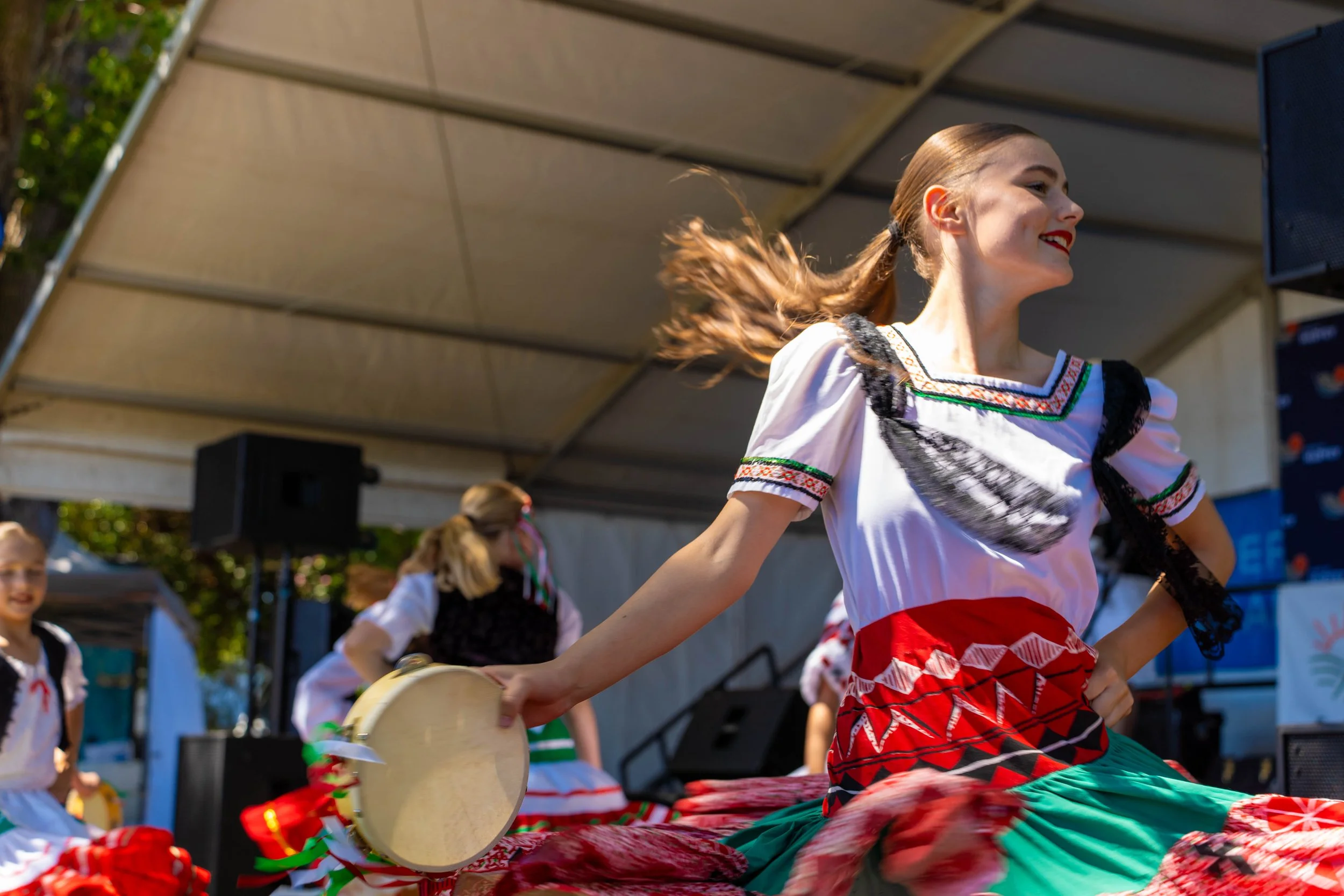 Young woman in traditional folk dress dancing on stage during cultural performance, with other dancers in similar attire in the background.