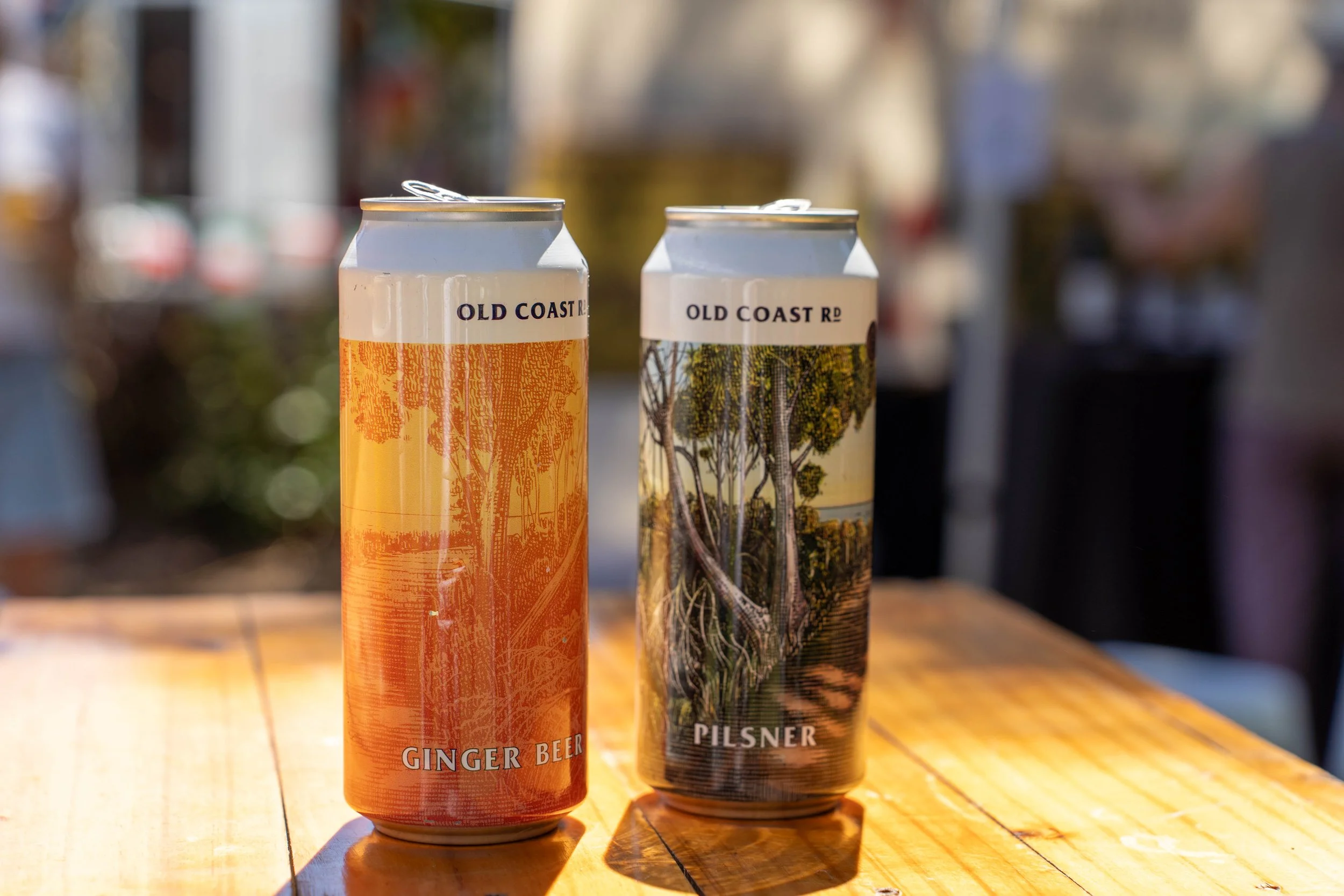 Two cans of beer on a wooden table outdoors with blurred background, one labeled Ginger Beer and the other labeled Pilsner, both from Old Coast Rd.