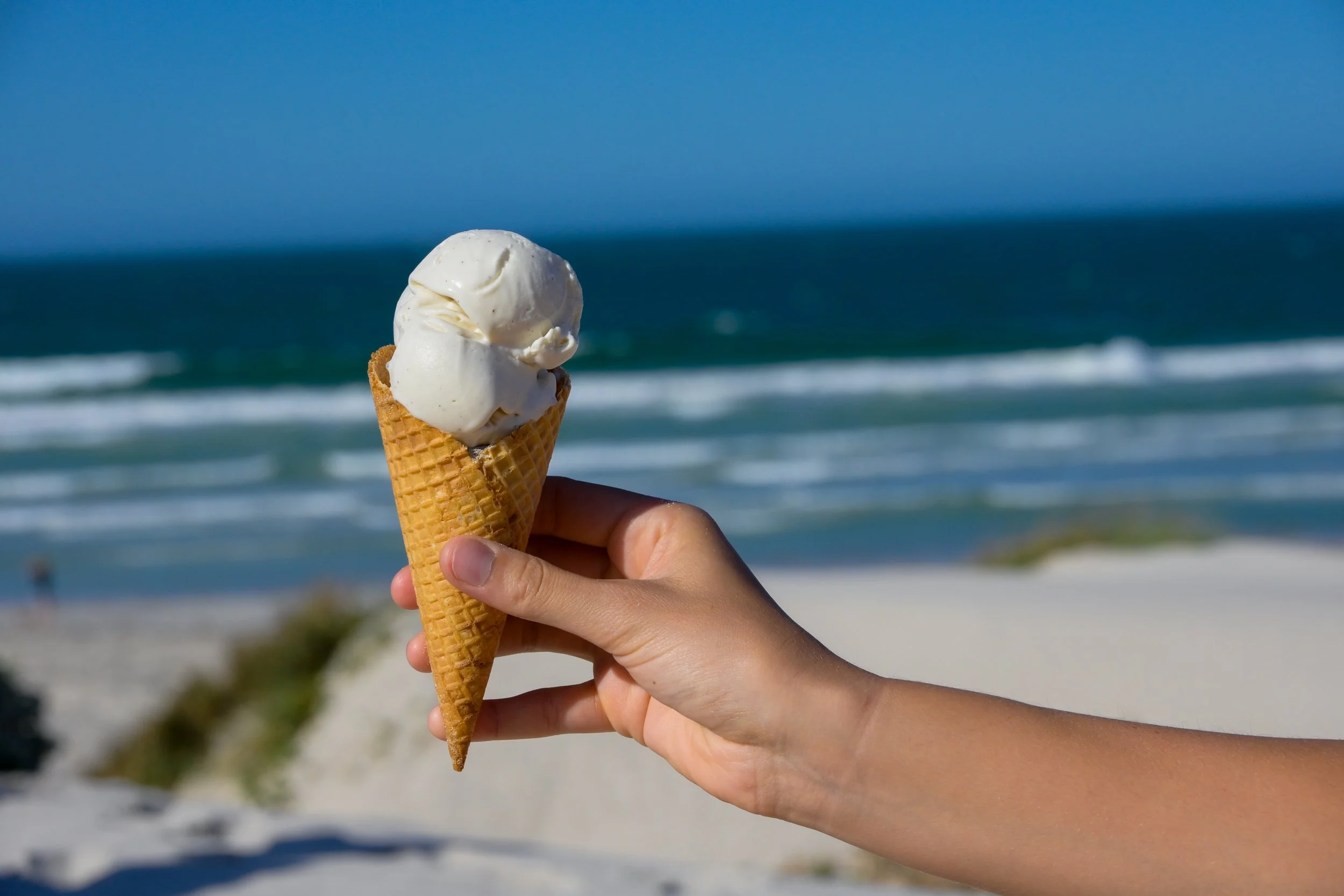 A bottle of buffalo milk, a dollop of cheese with a sprig of rosemary on a plate, a bowl of cherry tomatoes, and a person's hands reaching for the tomatoes on a green and white striped tablecloth outdoors.