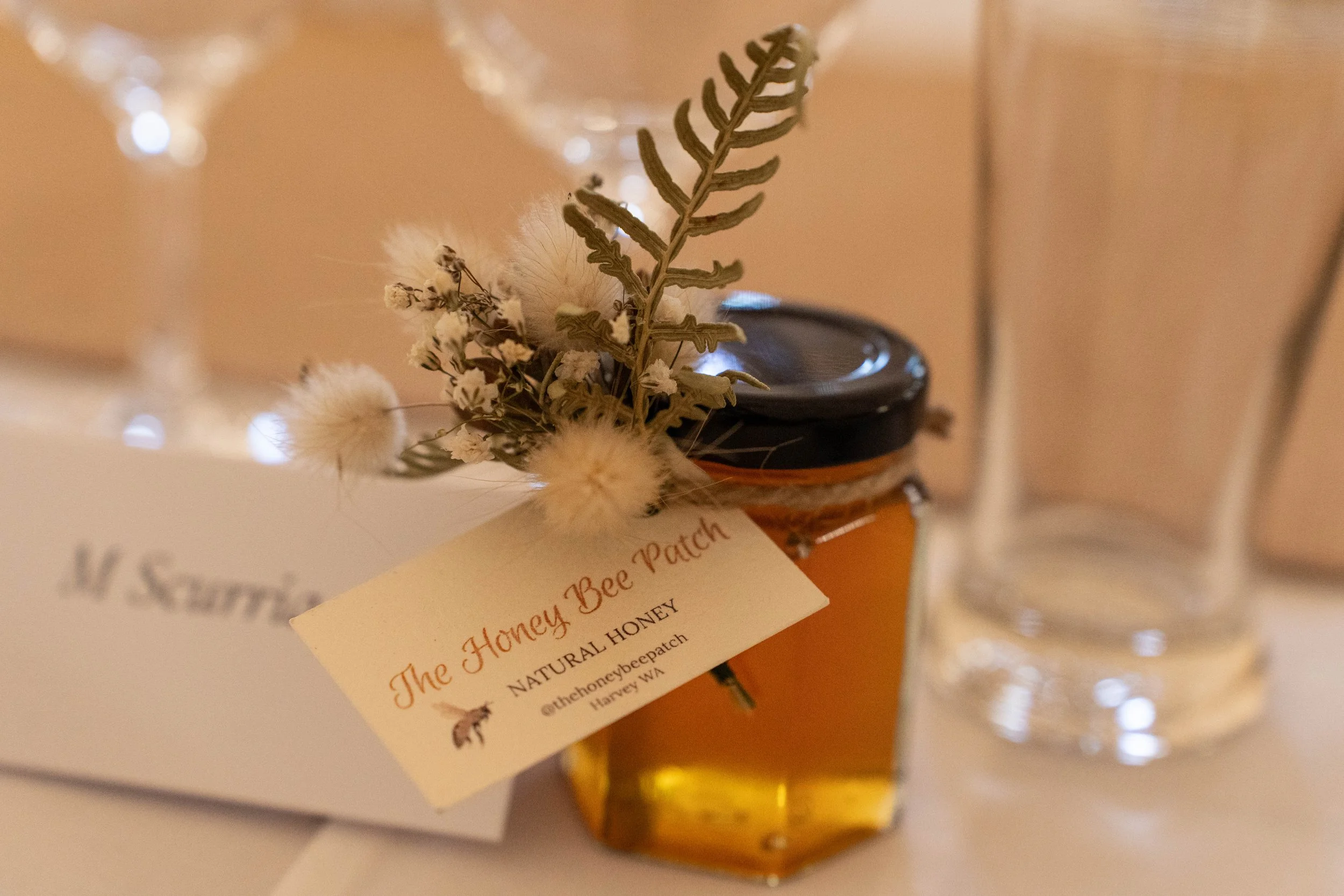 A jar of honey labeled 'The Honey Bee Patch, Natural Honey,' with a small bouquet of dried flowers attached, placed on a table with a clear glass and a white box in the background.