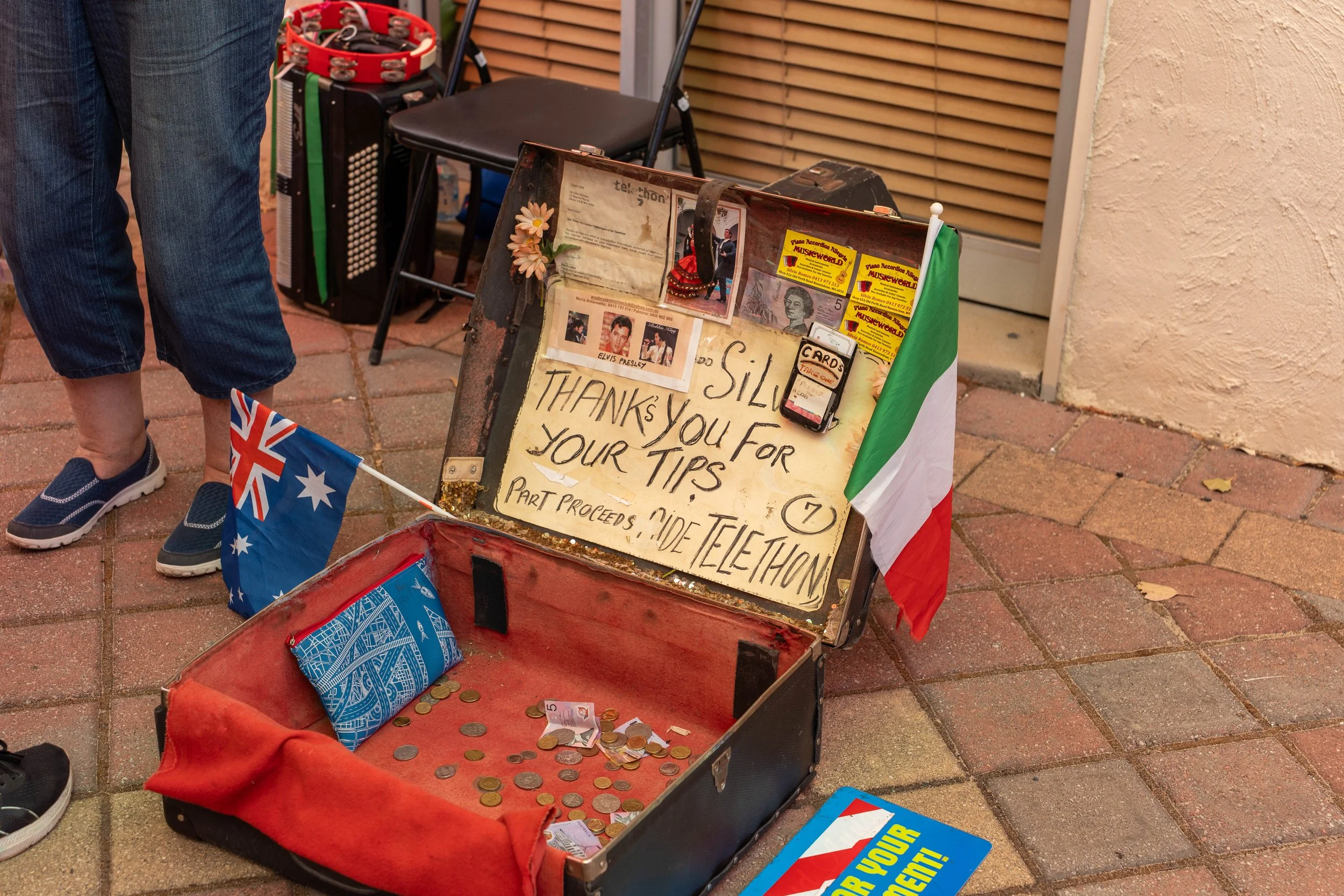 A small donation box with coins and bills, a sign thanking for tips, an Italian flag, and promotional materials on a folding display stand, with a person standing nearby holding Australian flags.