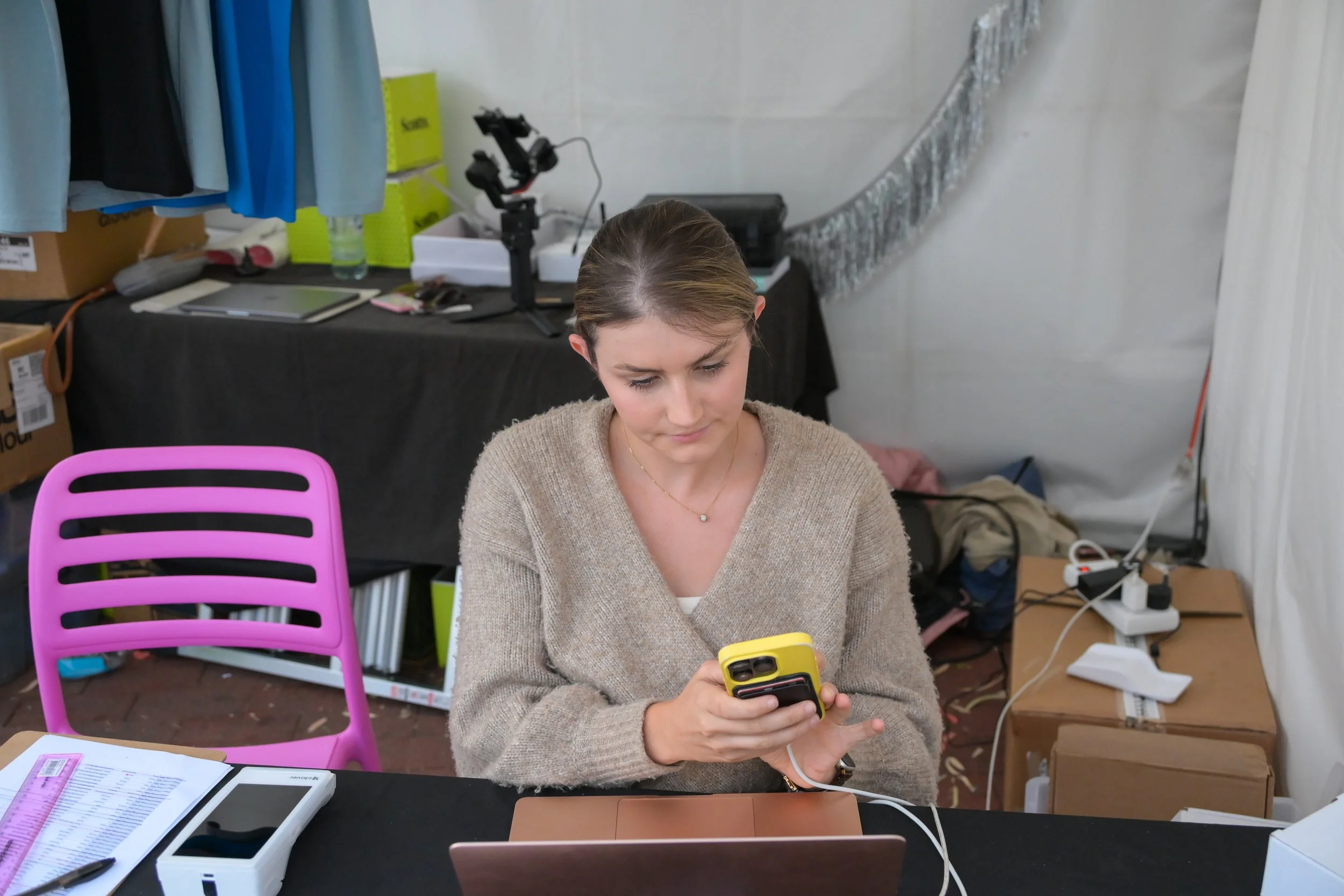A woman sitting at a desk, looking at her smartphone, with a laptop in front of her in a workspace or office setting.