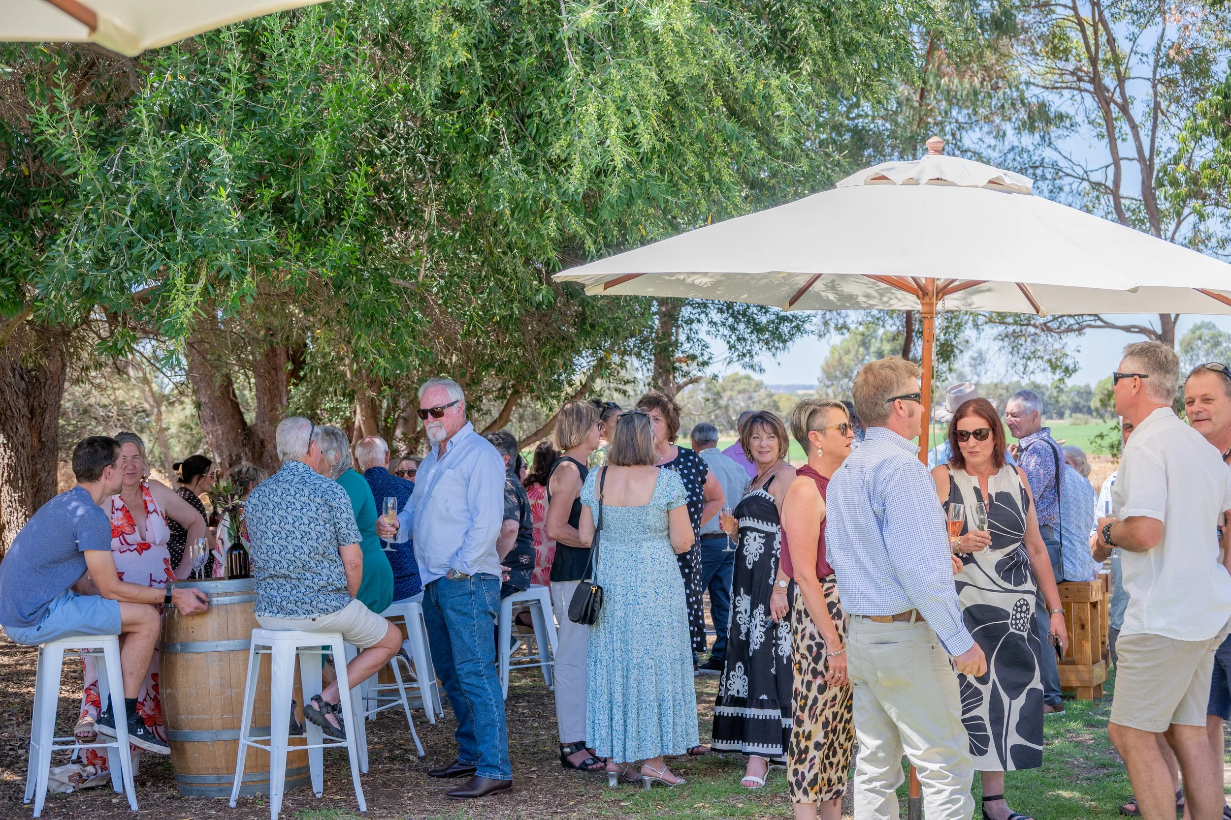 People gathered outdoors under large white umbrellas, socializing and enjoying drinks at an event on a sunny day with green trees in the background.