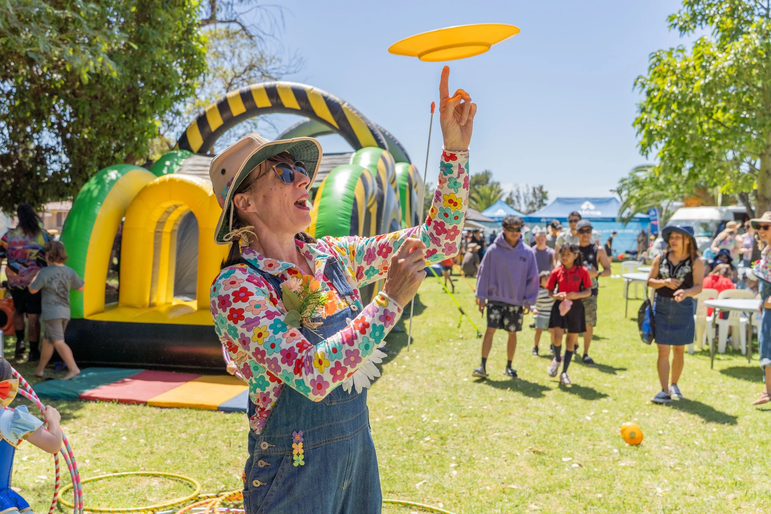 A woman in a floral shirt, overalls, and a beige sun hat is spinning a yellow flying disc at an outdoor event. There are inflatable structures and people in the background on a sunny day.