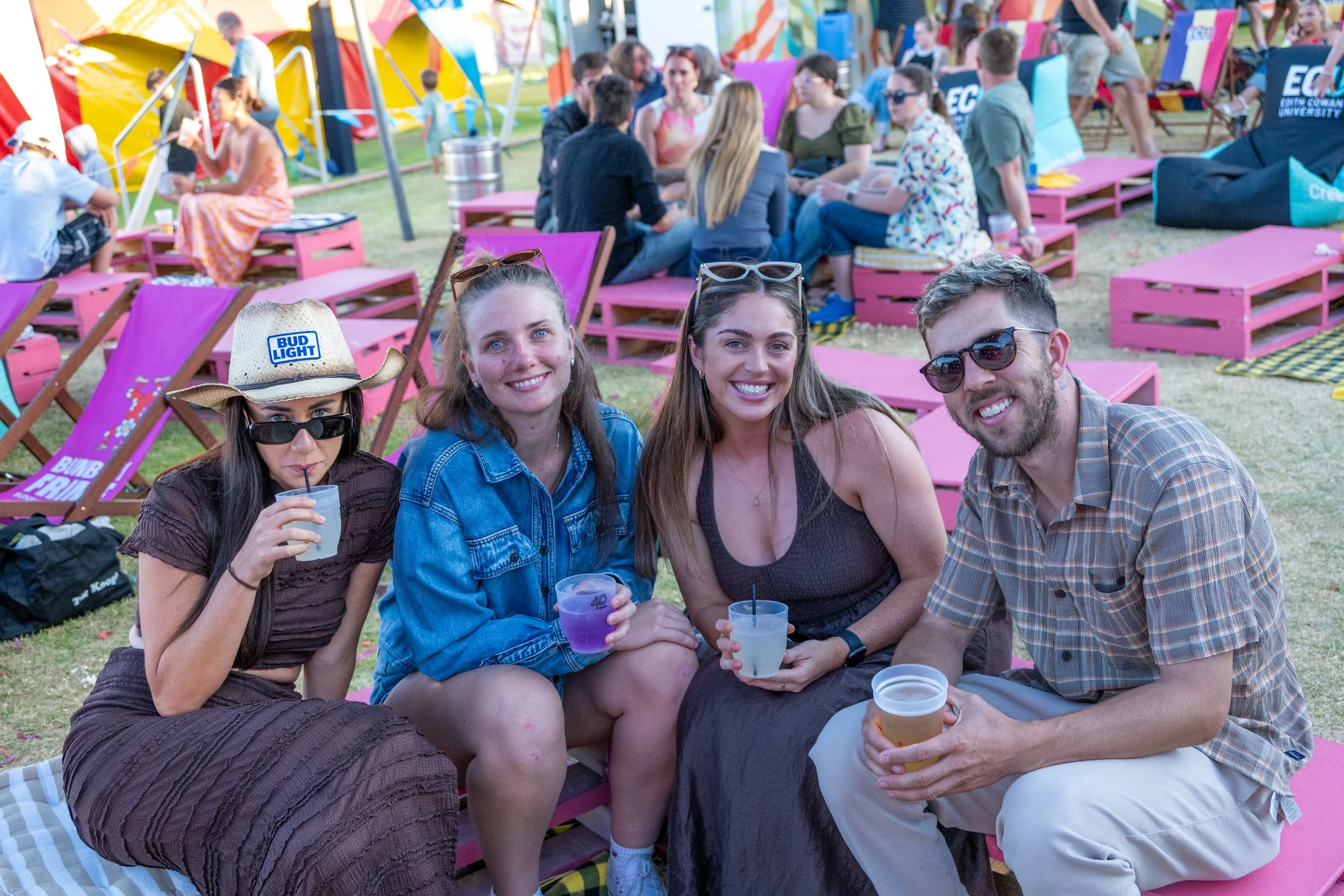 Four people sitting close together on pink benches at an outdoor event, smiling and holding drinks, with more people and colorful tents in the background.