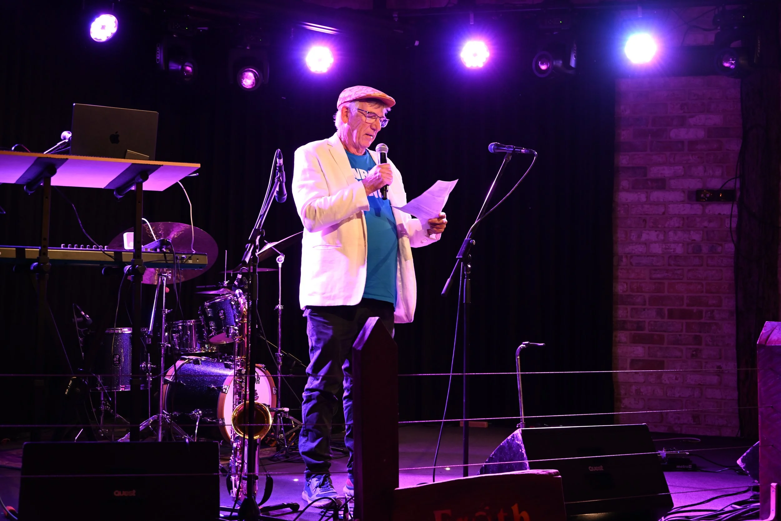 An older man wearing a white blazer, teal shirt, and cap standing on a stage with musical instruments, reading from a paper into a microphone under purple stage lights.