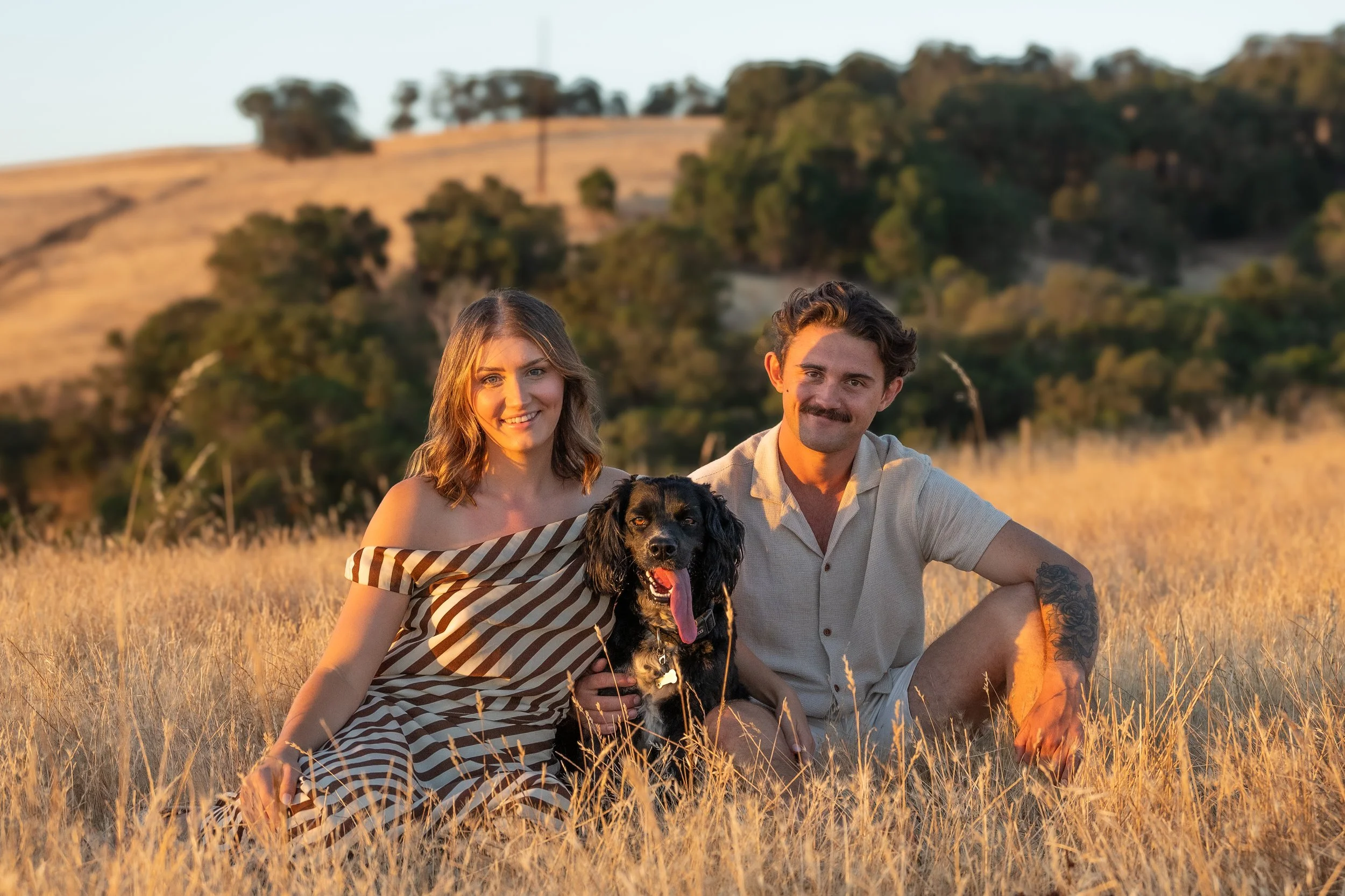 A smiling young woman, a smiling young man, and a black dog with floppy ears sitting in a field of dry grass during sunset.