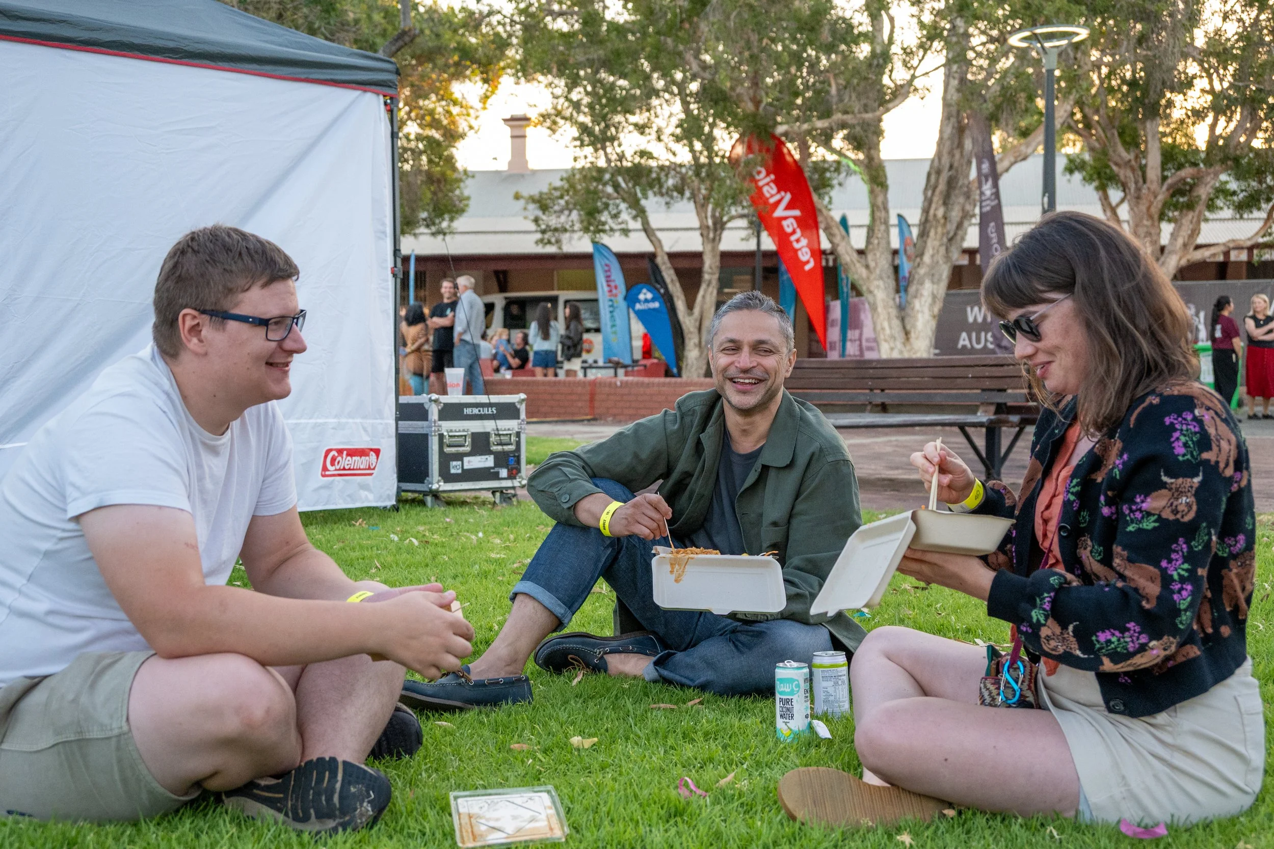 Three people sitting on grass at outdoor event, eating food, smiling and engaging in conversation, with flags and tents in the background.