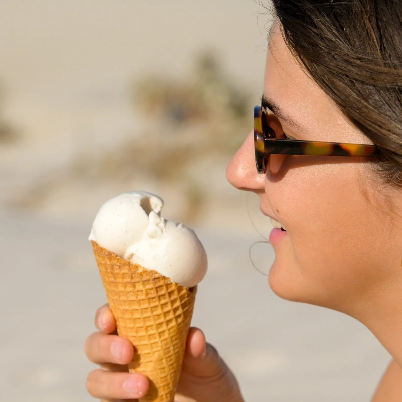 A woman wearing tortoise-shell sunglasses holding and about to eat a vanilla ice cream cone outdoors.