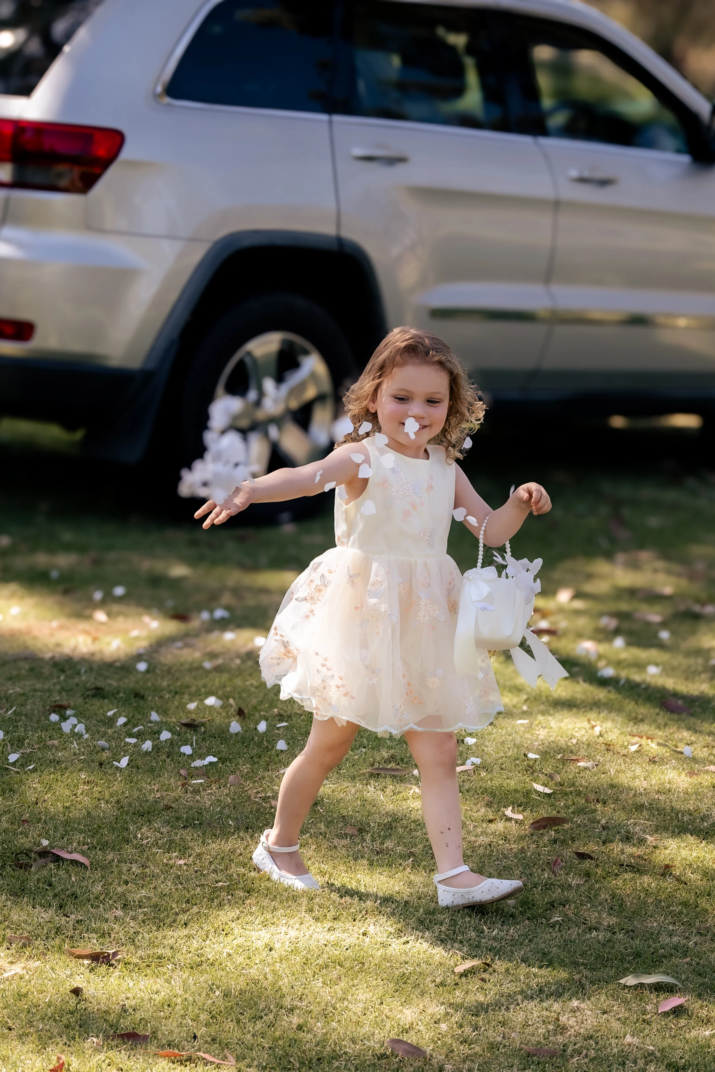 Young girl in a light-colored dress with floral details, holding a white basket, playing outside on grassy ground with fallen petals, near a parked silver SUV.