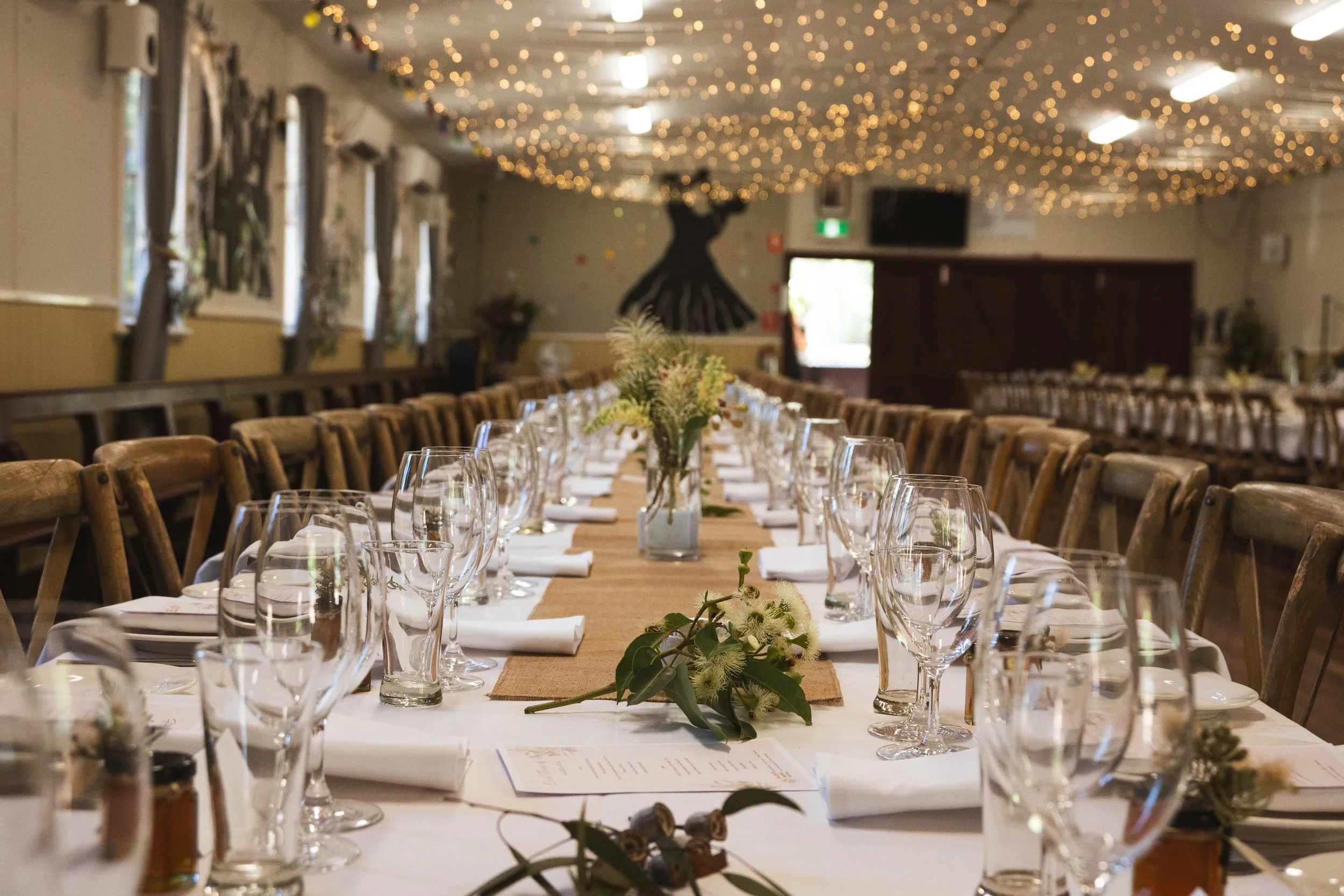 Decorated banquet table set for a celebration or event with wine glasses, white napkins, flowers, and menus, in a festive hall with string lights and wall decorations.