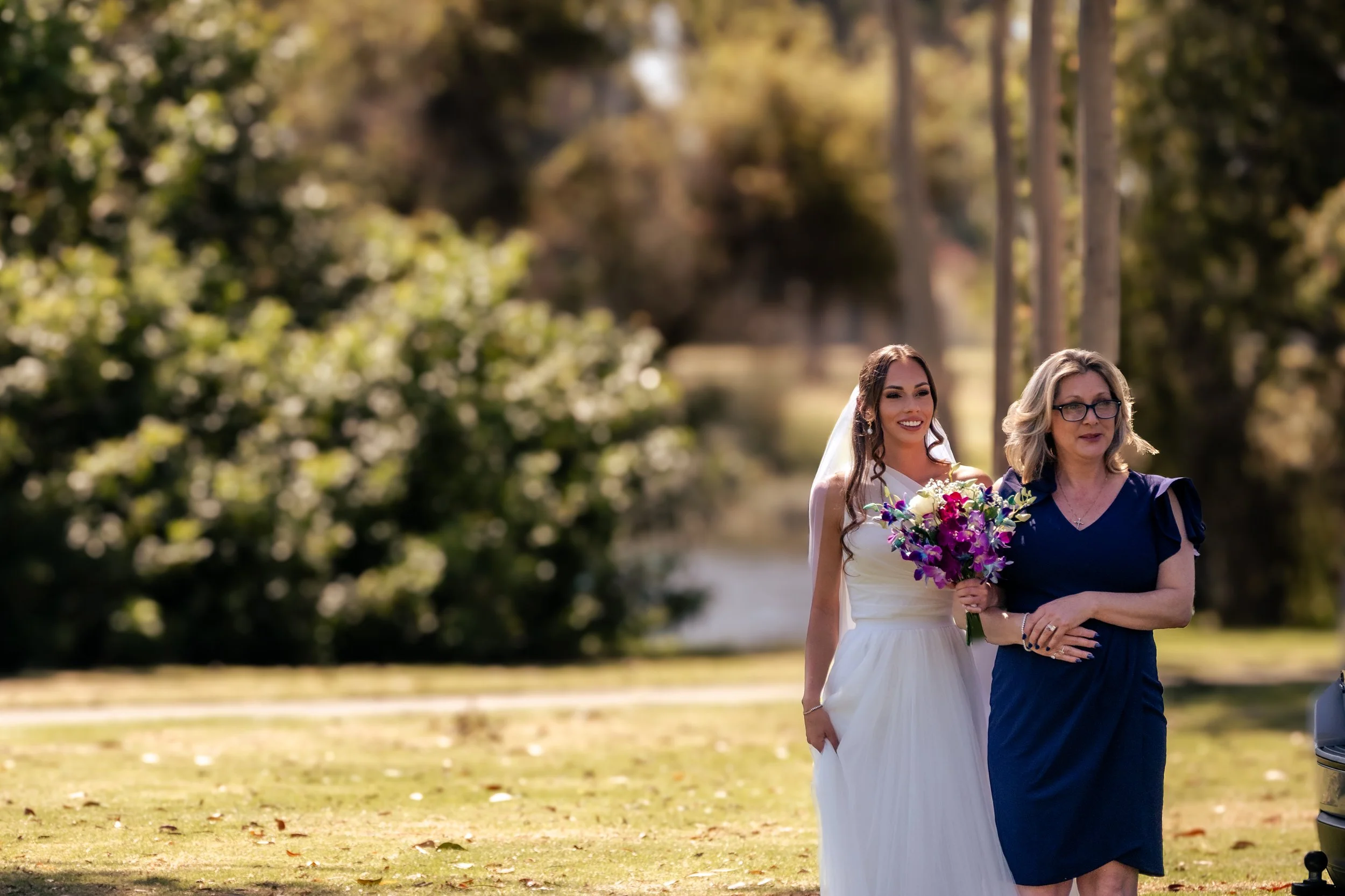 A bride in a white wedding dress is walking beside a woman in a blue dress with glasses, who appears to be walking her down the aisle outdoors in a park or garden with trees and grass.