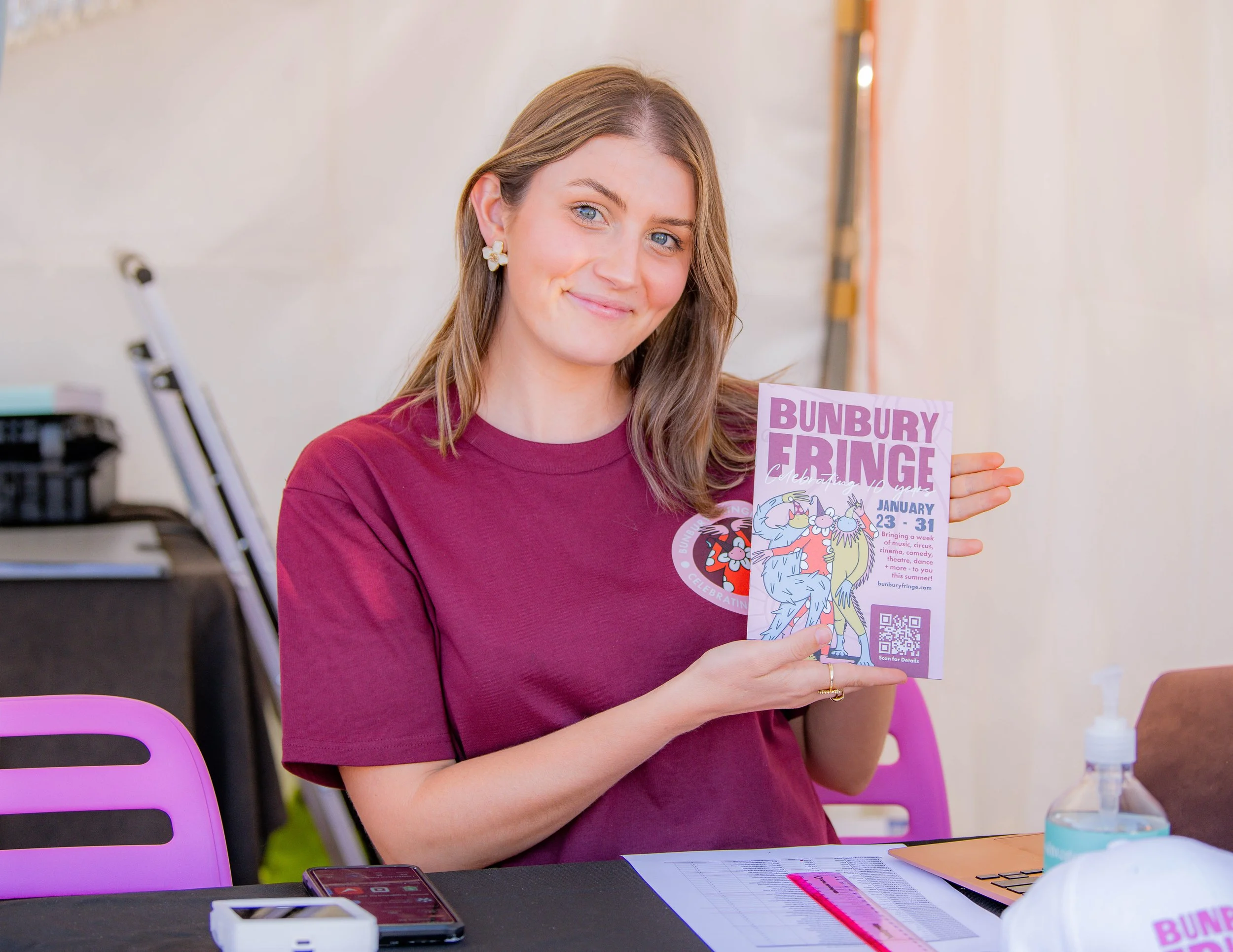 A woman with shoulder-length brown hair, wearing a burgundy t-shirt with Bunbury Fringe logo, smiling and holding a colorful event flyer that says 'Bunbury Fringe' with dates January 23-31, and has a QR code. She is sitting at a table with a pink chair, a smartphone, papers, a hand sanitizer, and a white helmet.