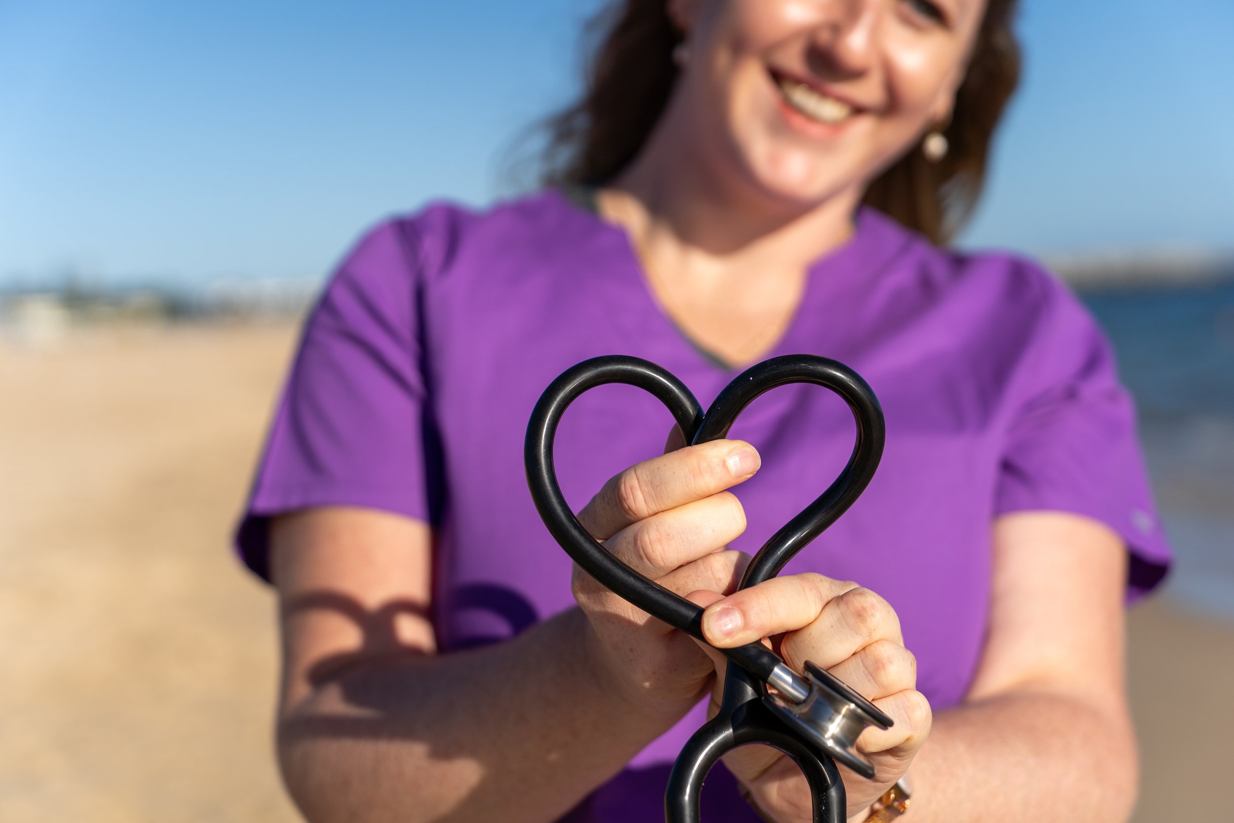 A smiling woman in a purple shirt holding a black stethoscope formed into a heart shape on a beach with a blue sky and ocean in the background.