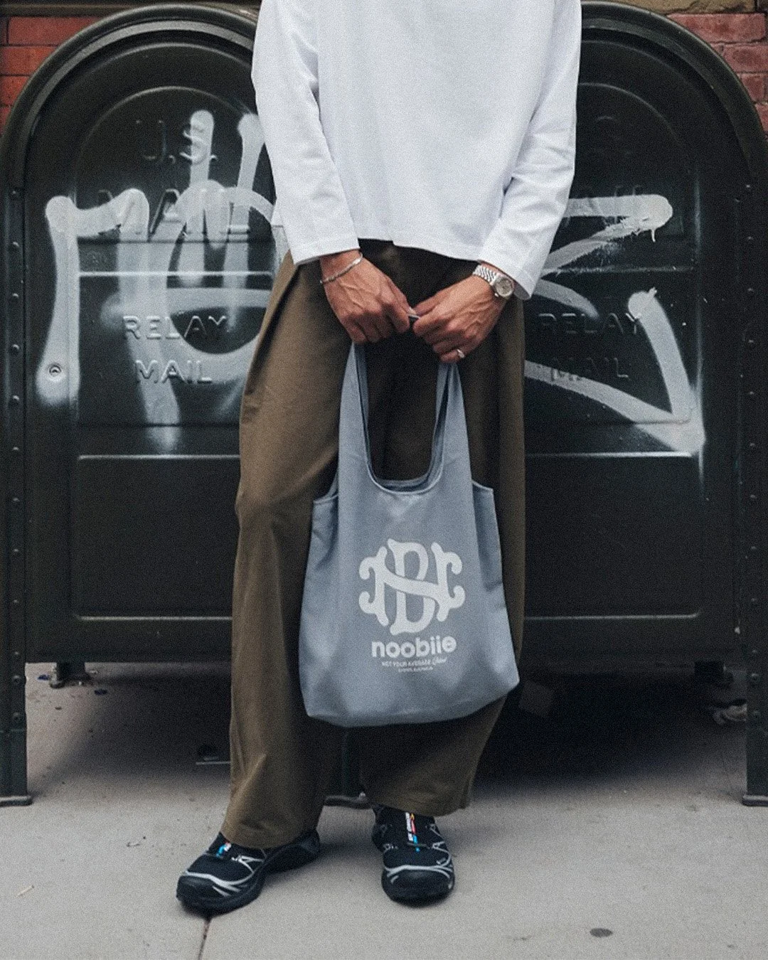 Person standing in front of a black mailbox, holding a gray tote bag with the word 'noobite' on it. The individual is wearing a white long-sleeved shirt, brown pants, black sneakers, a watch, and a bracelet.