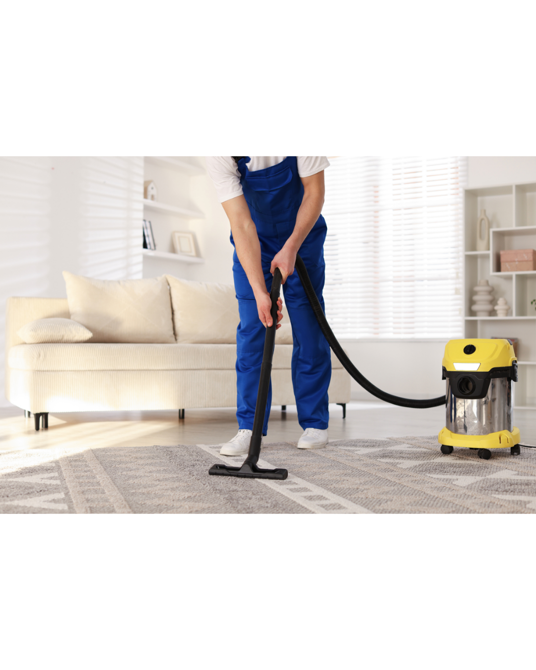Person in a white shirt and blue overalls vacuuming carpet in a living room with a yellow and black vacuum cleaner.
