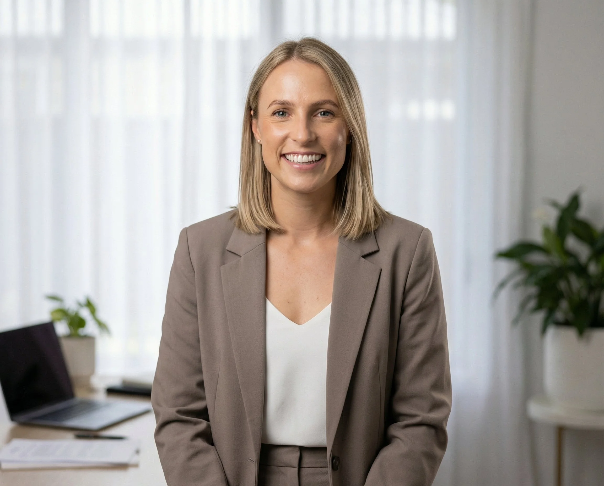 A smiling surgeon with blonde hair dressed in a beige suit, standing in a bright office with a laptop and papers on a desk and green plants in the background.
