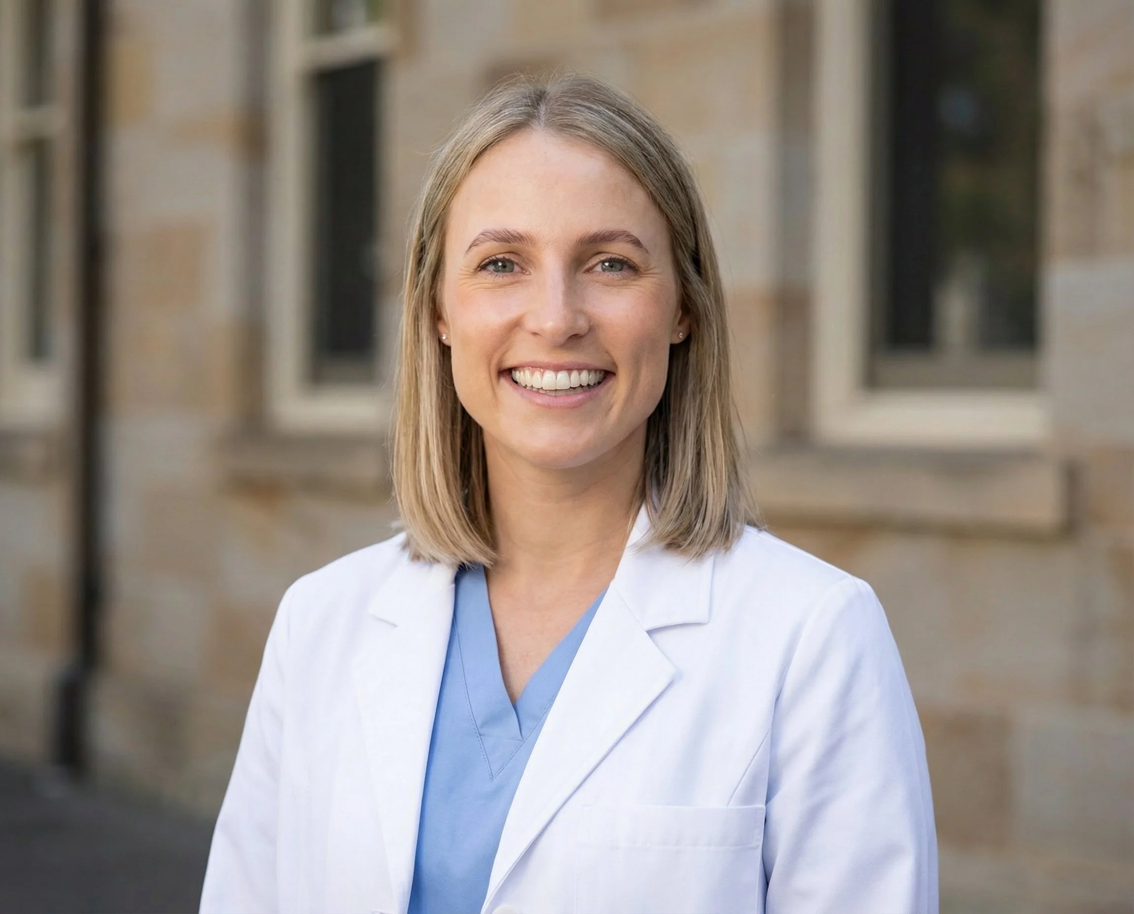 A smiling female surgeon healthcare professional in a white coat and blue scrubs standing outdoors in front of a brick building.