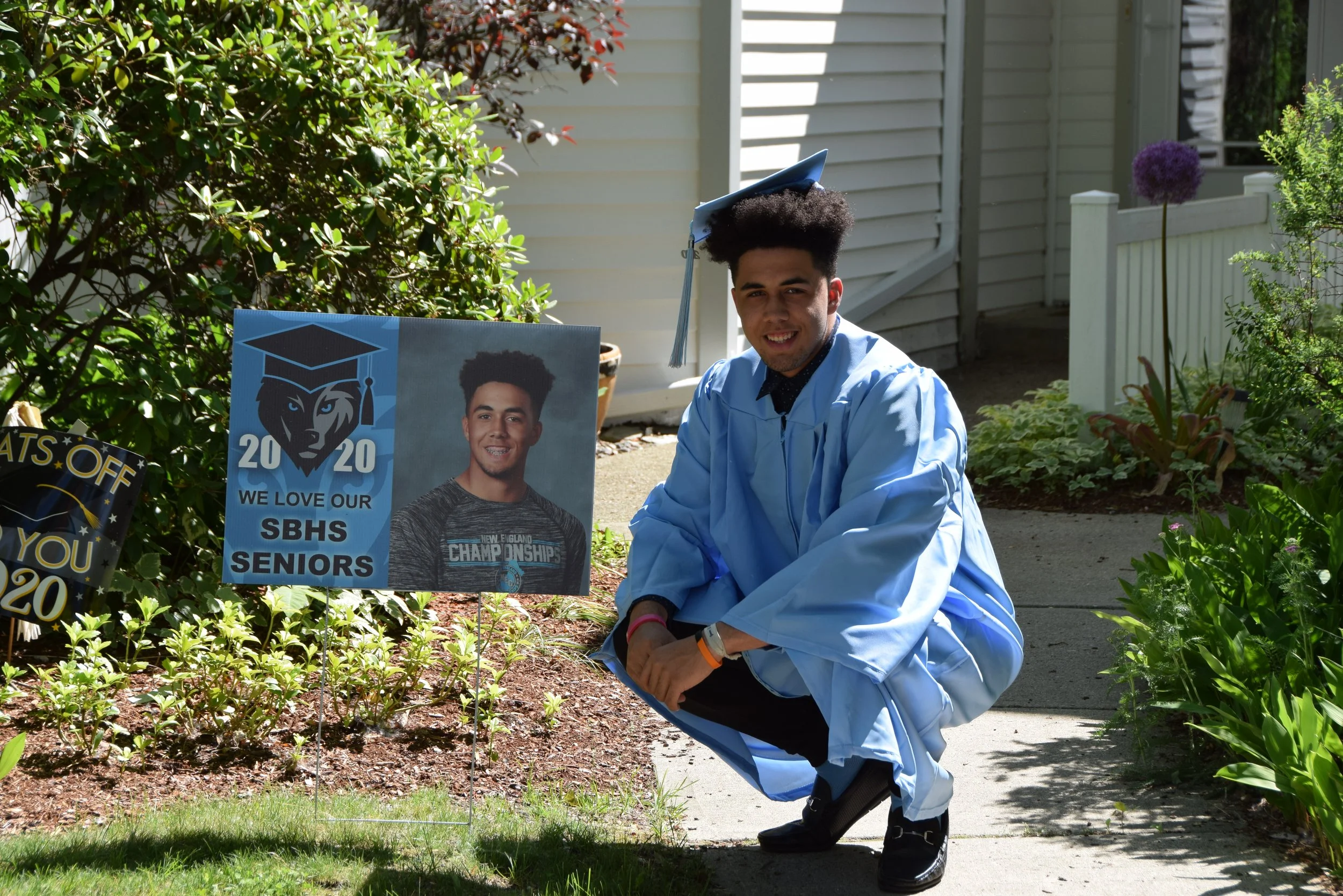 Javen Sears graduating in 2020 in a light blue graduation gown and cap, squatting on a sidewalk, smiling. Behind him, a sign with his photo, a graduation cap logo, and text expressing love for his senior class.