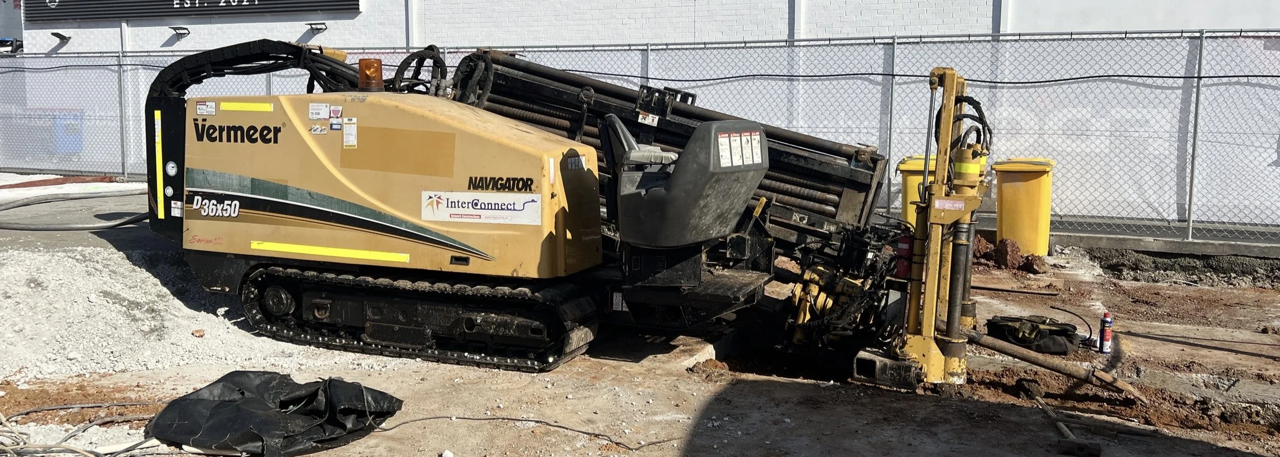 A Vermeer Navigator D36x50 drilling machine at a construction site, with a chain-like track for movement, a control panel, and a mounted drilling apparatus, surrounded by construction materials, a chain-link fence, and safety equipment.
