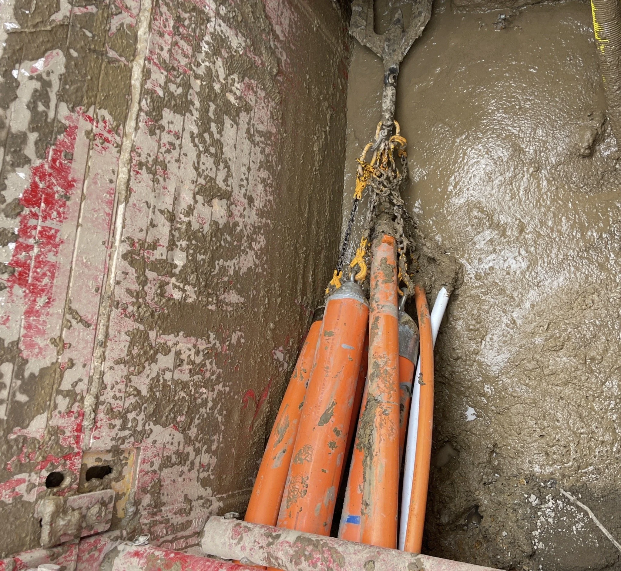 Orange and white pipes and chains in a construction site with muddy surroundings and a partially exposed wall.