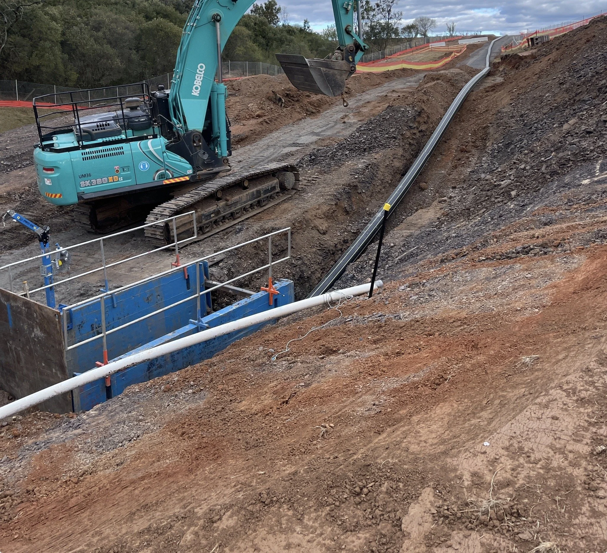 Construction site with a blue excavator working on laying underground pipes on a dirt hillside.