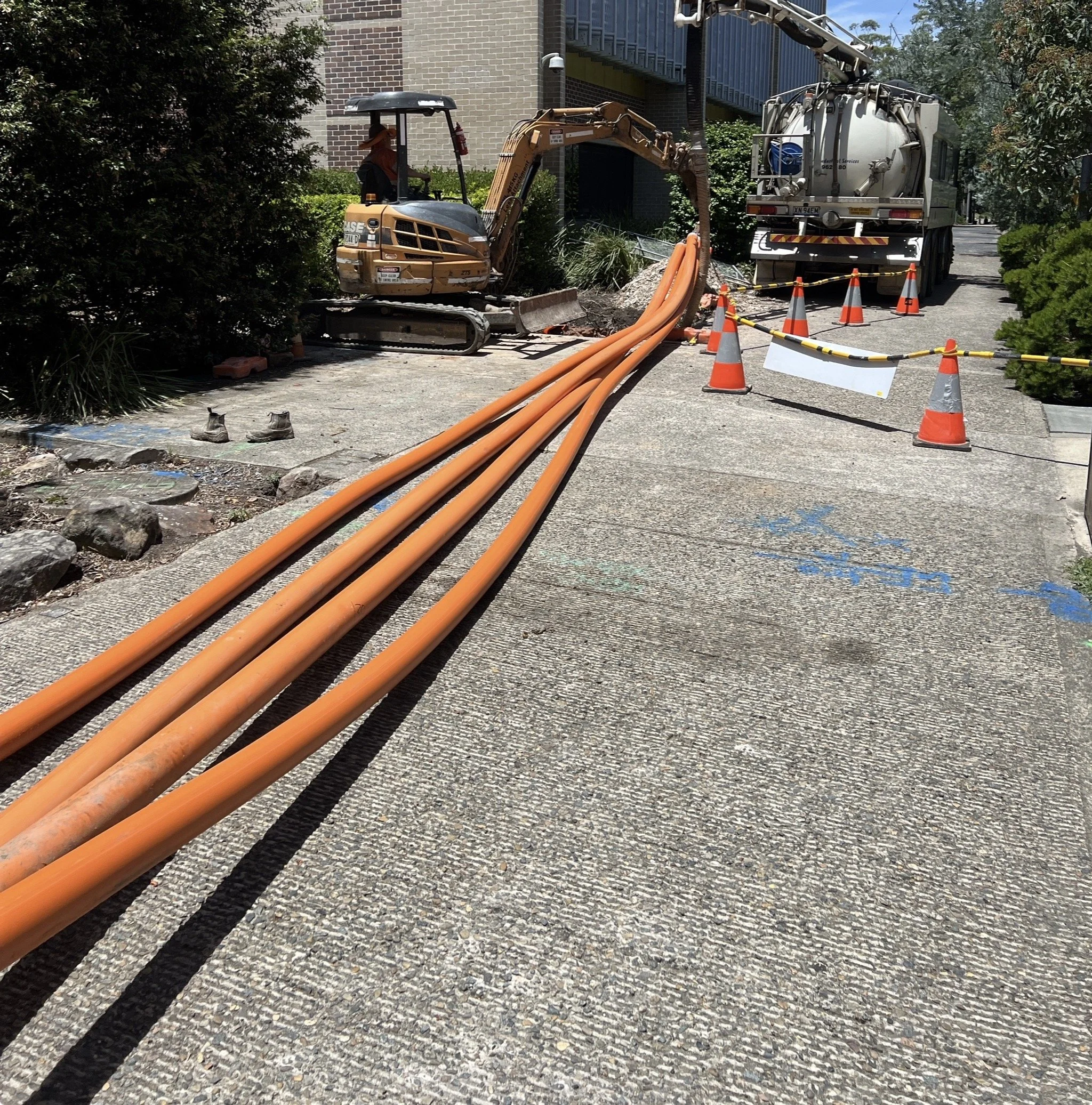 A construction site on a sidewalk showing several long orange cables, a small excavator, a construction worker, traffic cones, and a vacuum truck in the background.