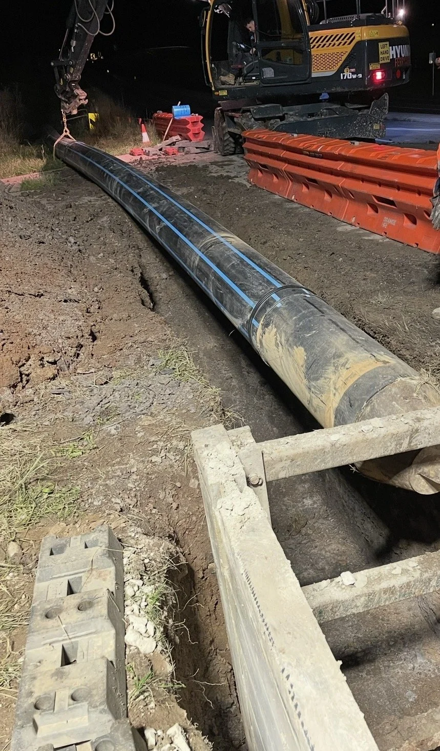 Construction site at night with a large pipe in a trench, a crane, and construction barriers.