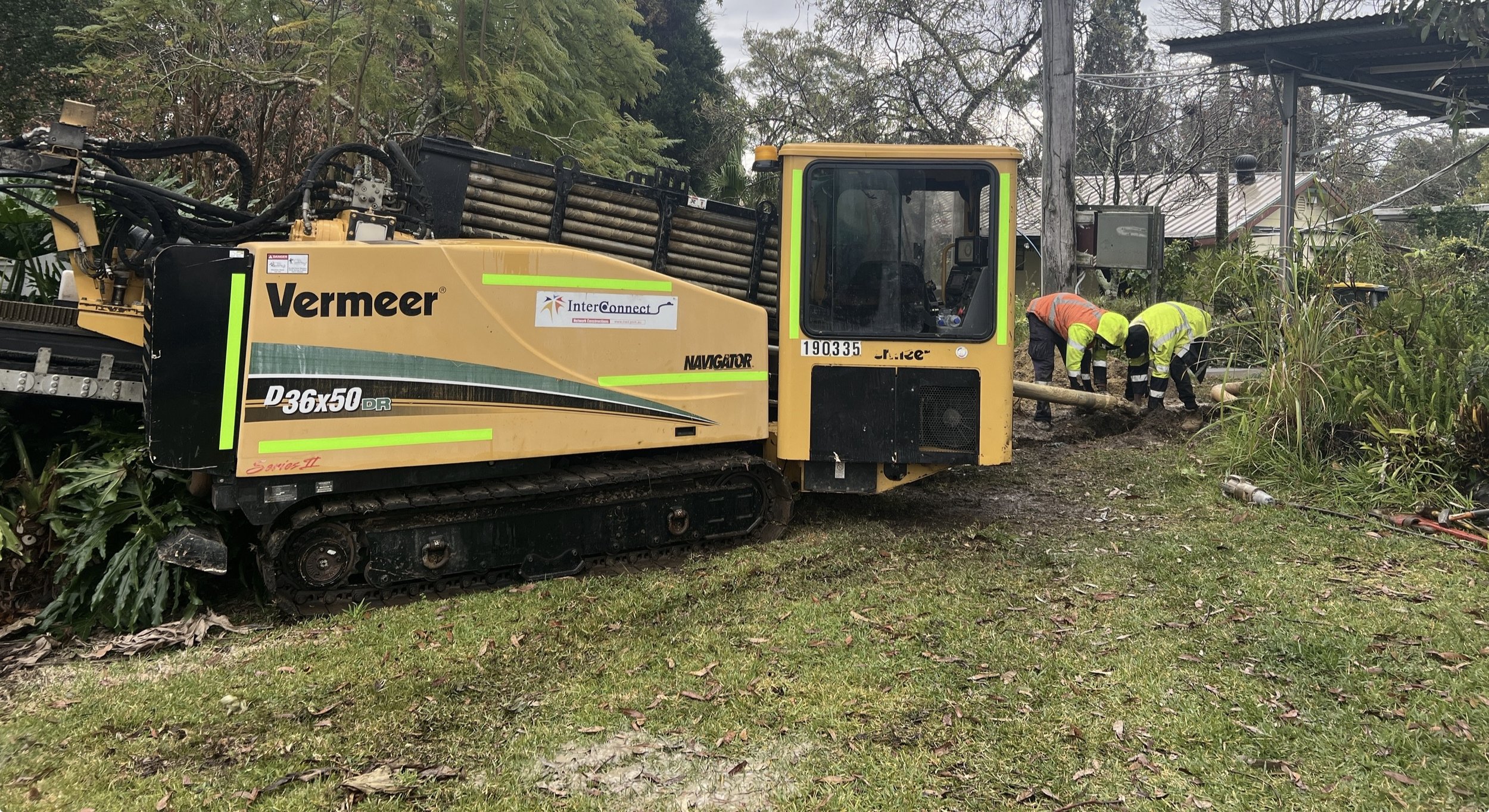 Construction workers in safety gear operating a Vermeer D36x50DR Navigator drill rig outdoors, with trees and a house in the background.