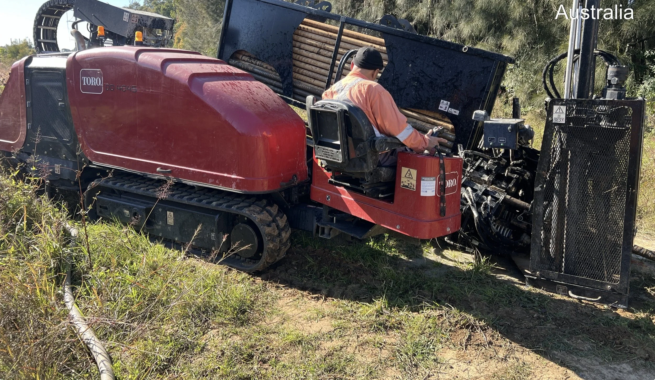 A worker operating a red Toro Dingo compact utility vehicle on grassy terrain in Australia, with a black attachment on the front used for trenching or digging. The worker is wearing an orange jacket and a black beanie, and is sitting in the operator'