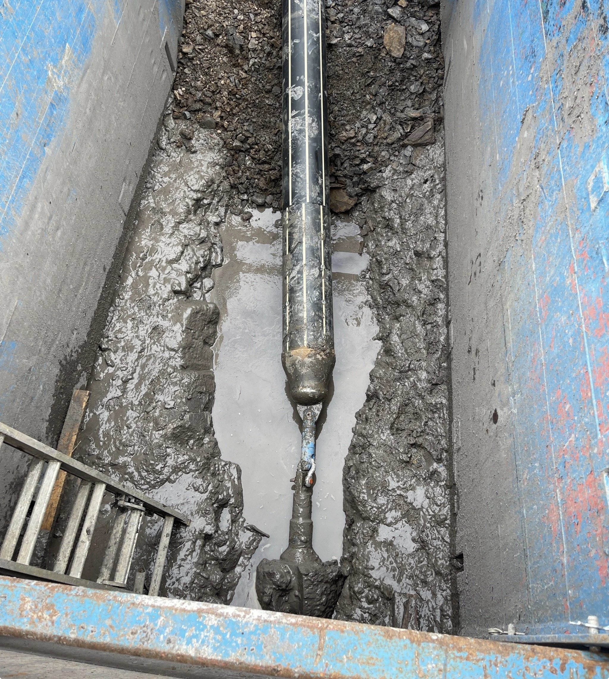 Construction site showing a large pipe in a trench with mud and water, surrounded by concrete walls with blue markings.