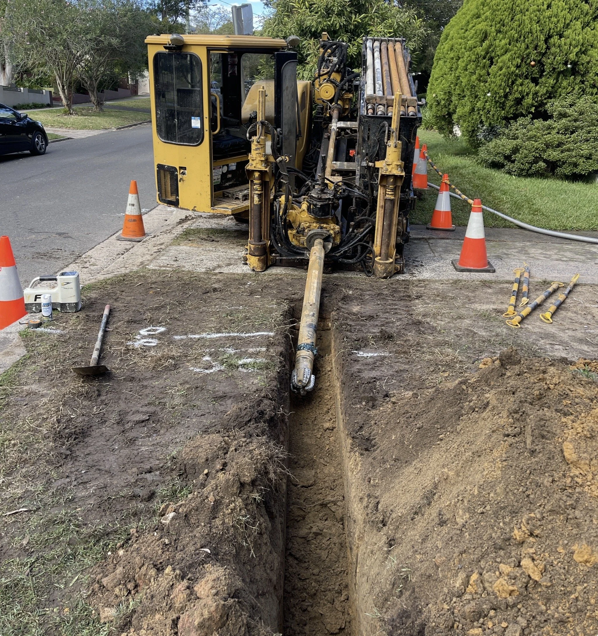 A construction site on a residential street with a large yellow and black drilling machine in the middle of a trench. Orange traffic cones surround the work area, and various tools and equipment are nearby.