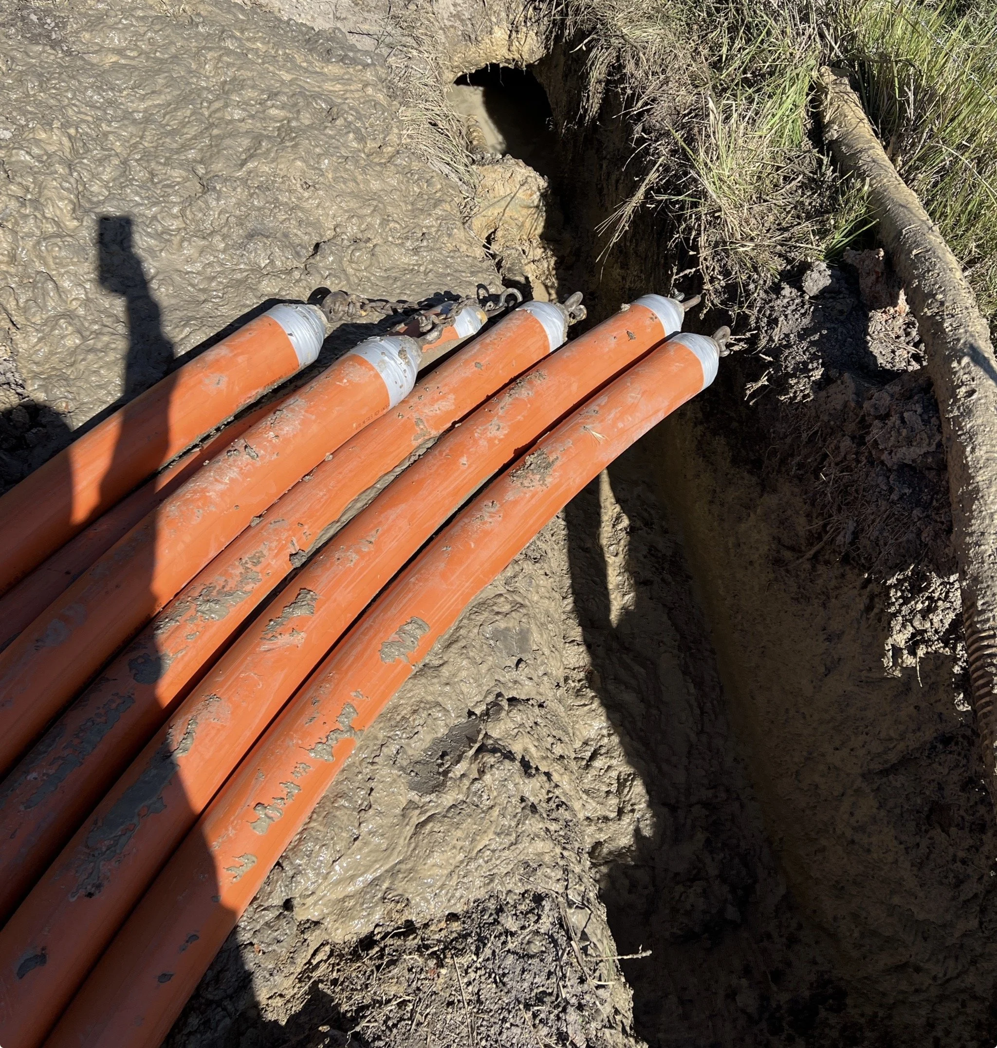 Several orange and silver utility pipes are laid in a trench in muddy soil, with some grass and a tree branch nearby.