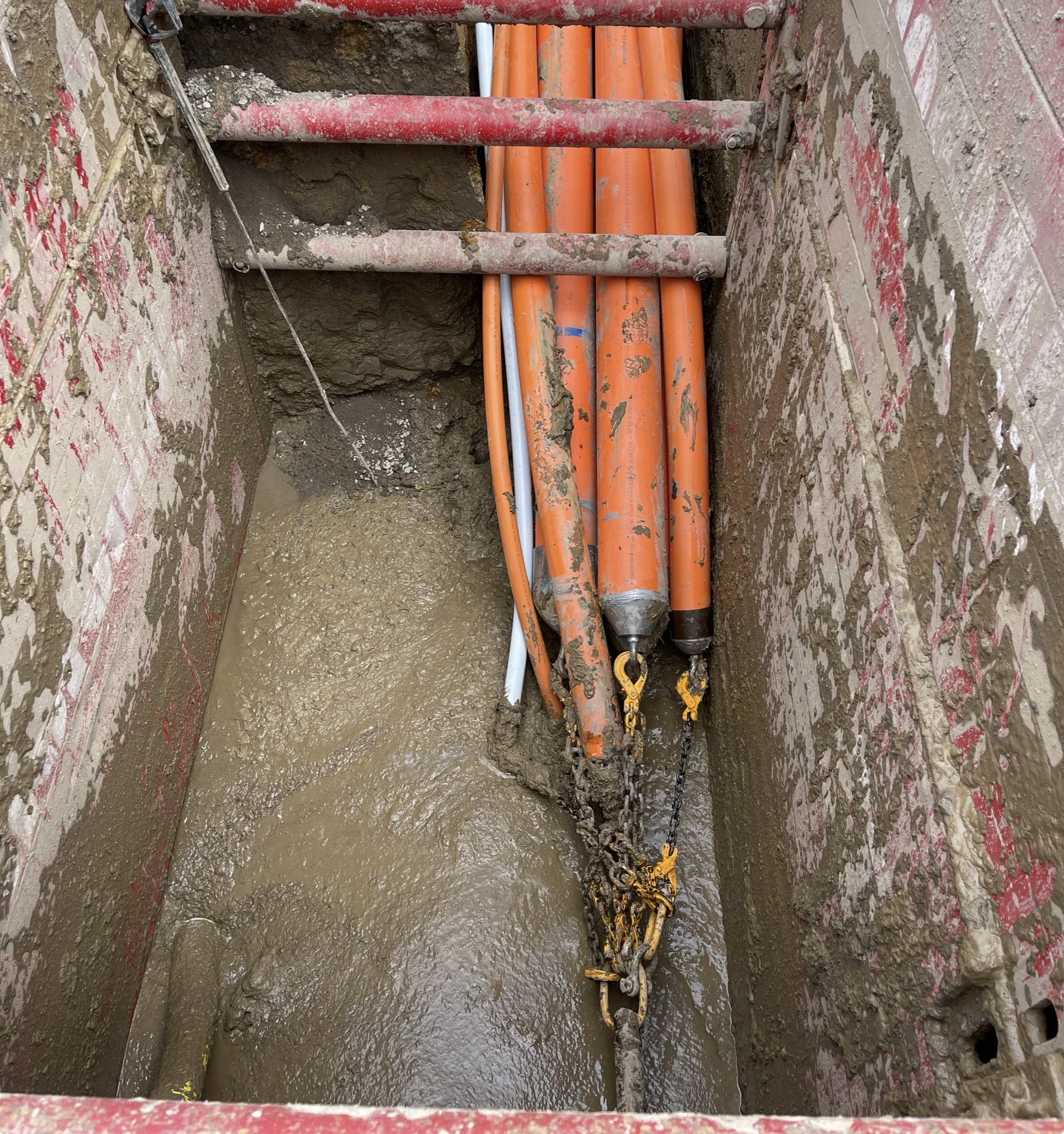 Construction site with orange jackhammers, chains, and muddy ground in a deep trench.