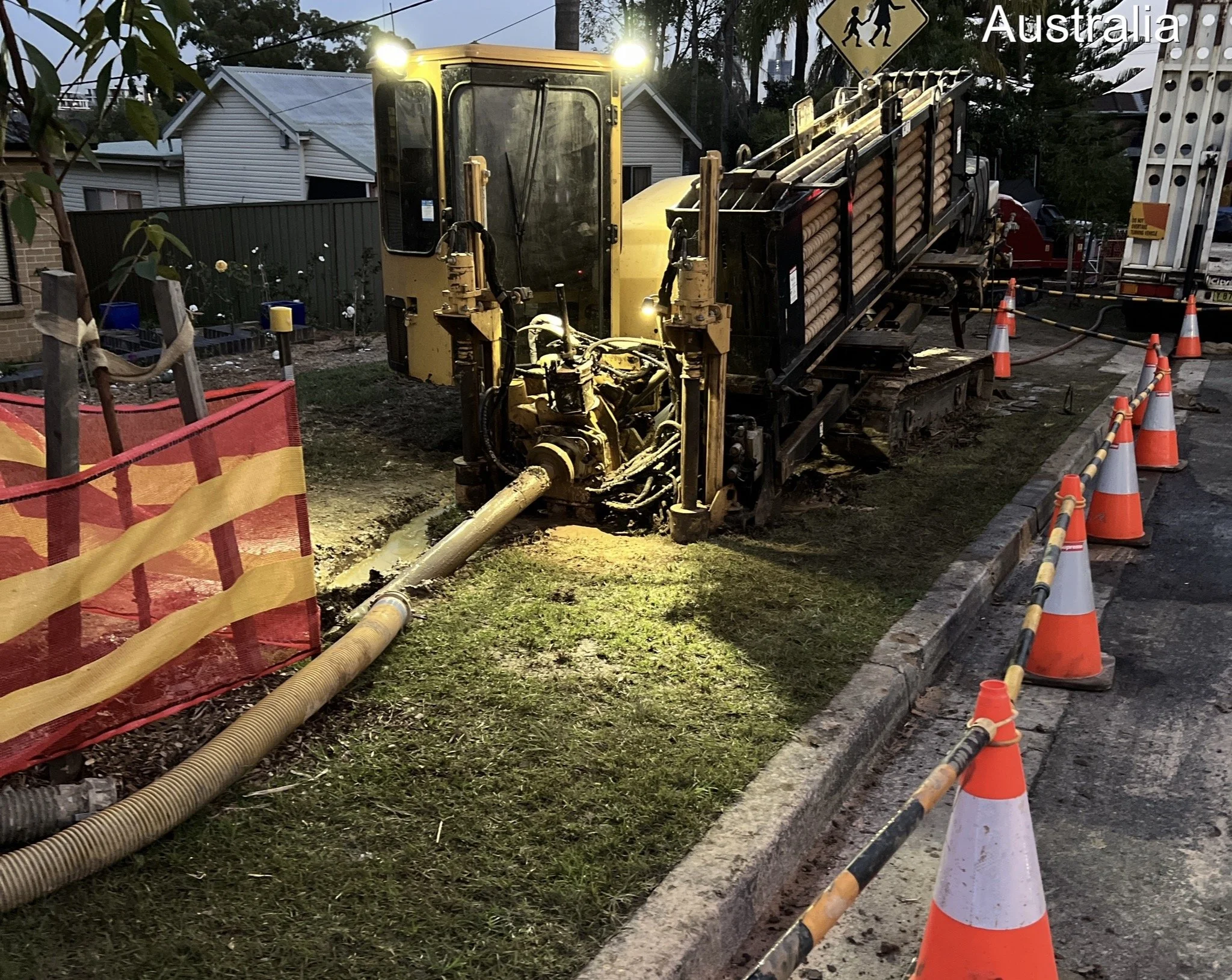 A construction worker is using a large machine to drill into the ground next to a sidewalk, with several orange and white traffic cones and a red barrier nearby. Residential houses are visible in the background, and the scene is lit with artificial l