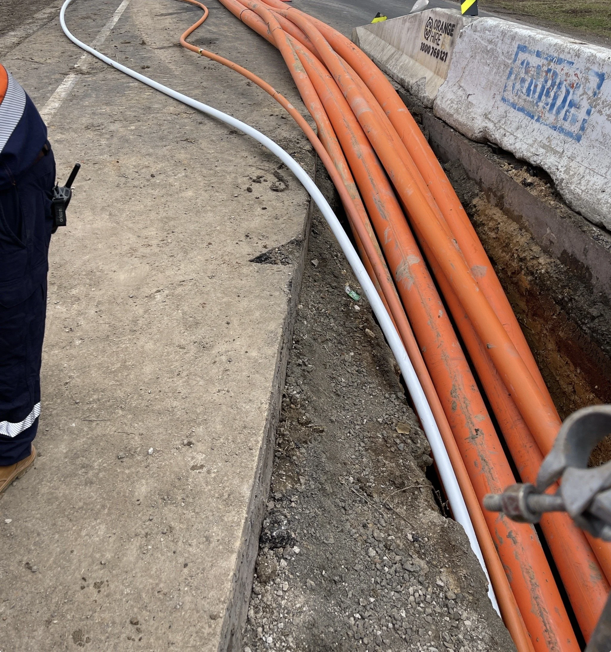 Construction site showing orange and white conduit pipes laid along the edge of a sidewalk under construction, with concrete barriers and safety barriers nearby.