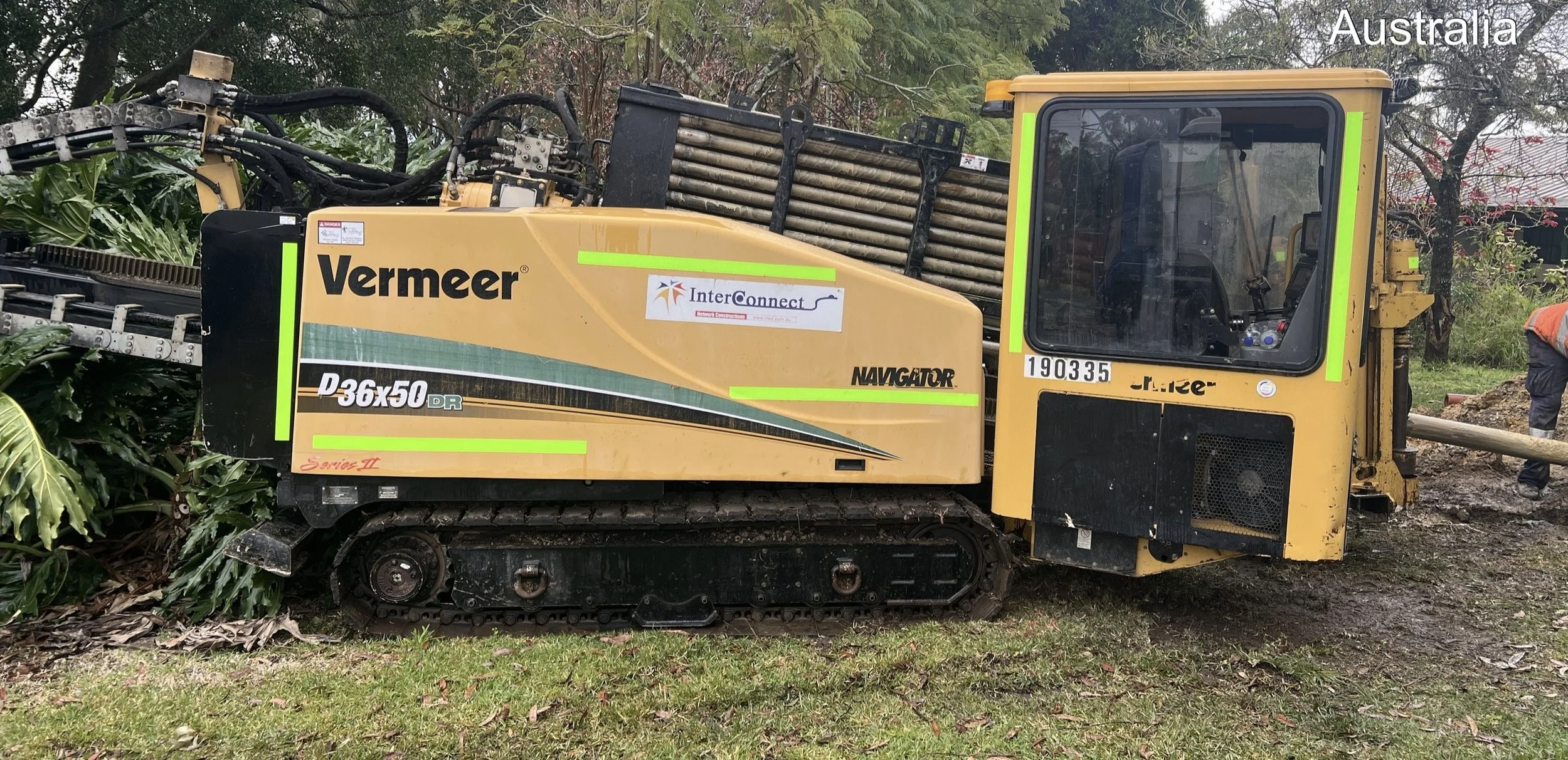 A yellow Vermeer D36x50 Navigator small construction excavator with black tracks, parked outdoors next to green plants and trees, with some construction equipment and a person in an orange vest nearby in Australia.