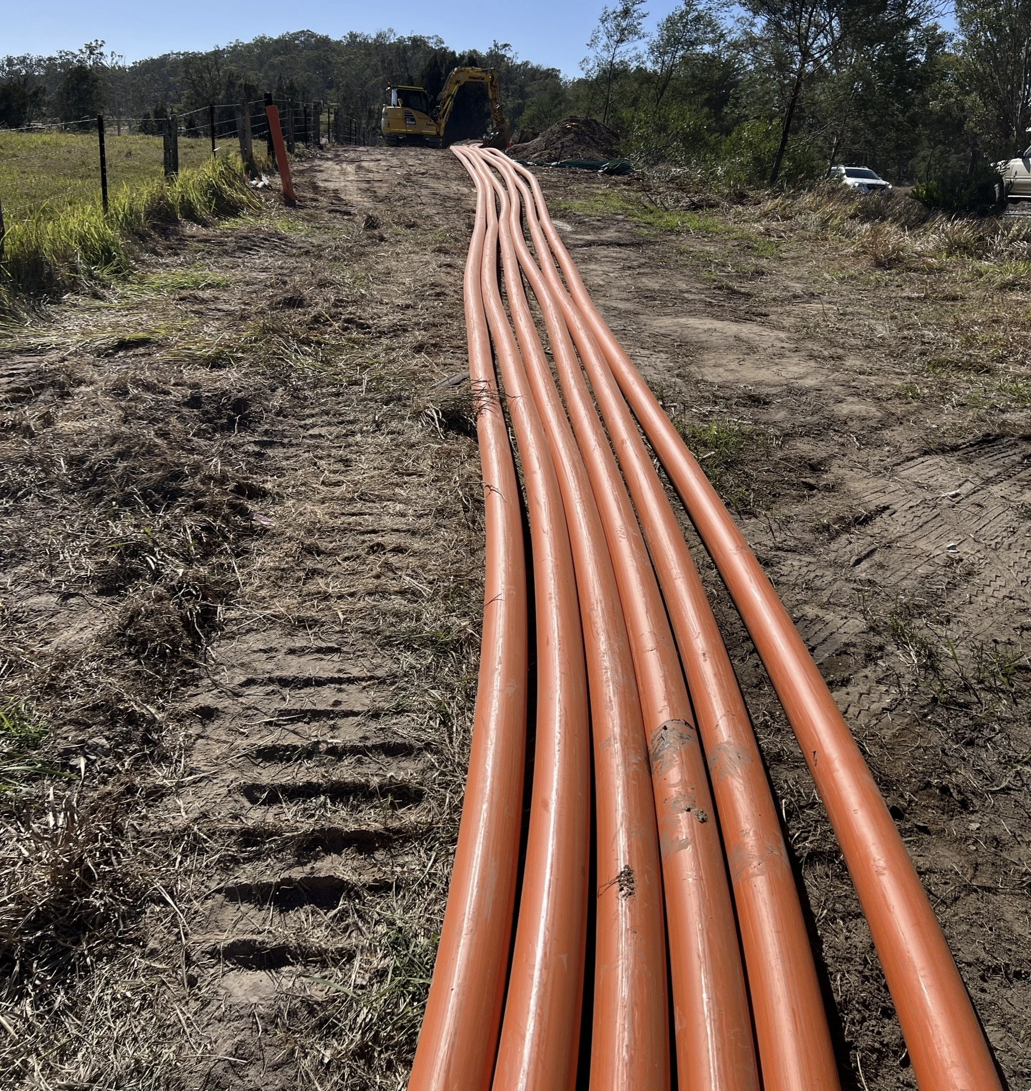 Multiple orange pipes laid out on a dirt path at a construction site, with a yellow excavator and trees in the background.