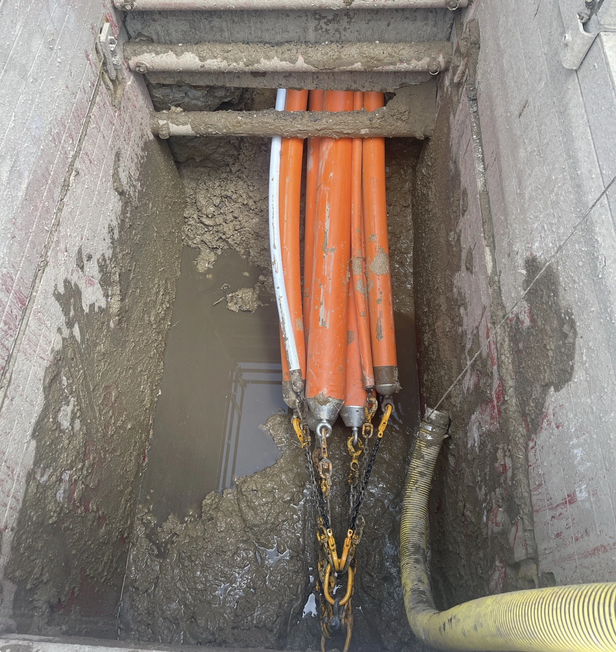 Construction site with orange and white pipes in a muddy, excavated trench.